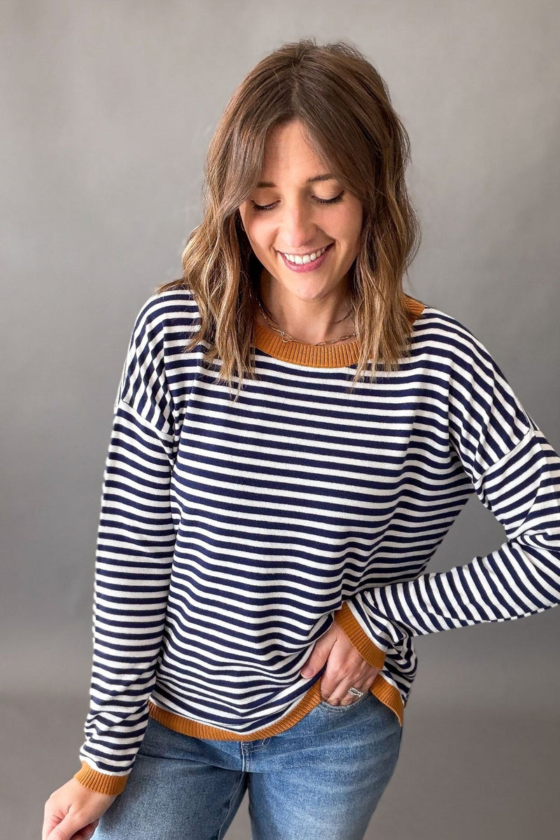 A woman wearing a navy and white horizontally striped long-sleeve sweater with a mustard yellow ribbed crew neck, cuffs, and hem, paired with blue jeans, against a neutral gray studio background.