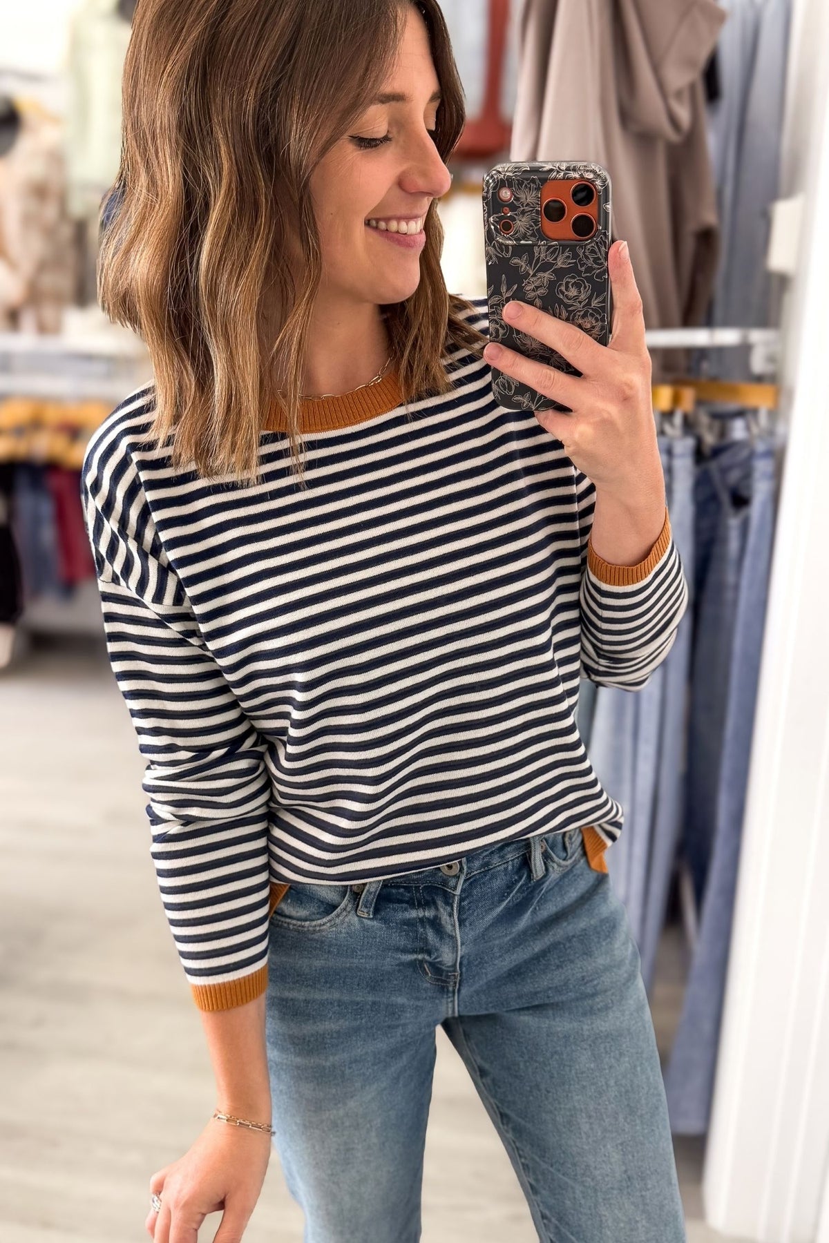 A woman takes a mirror selfie in a clothing store, wearing a black and white horizontally striped long-sleeve top with brown ribbed trim at the crew neck and cuffs, paired with blue jeans.