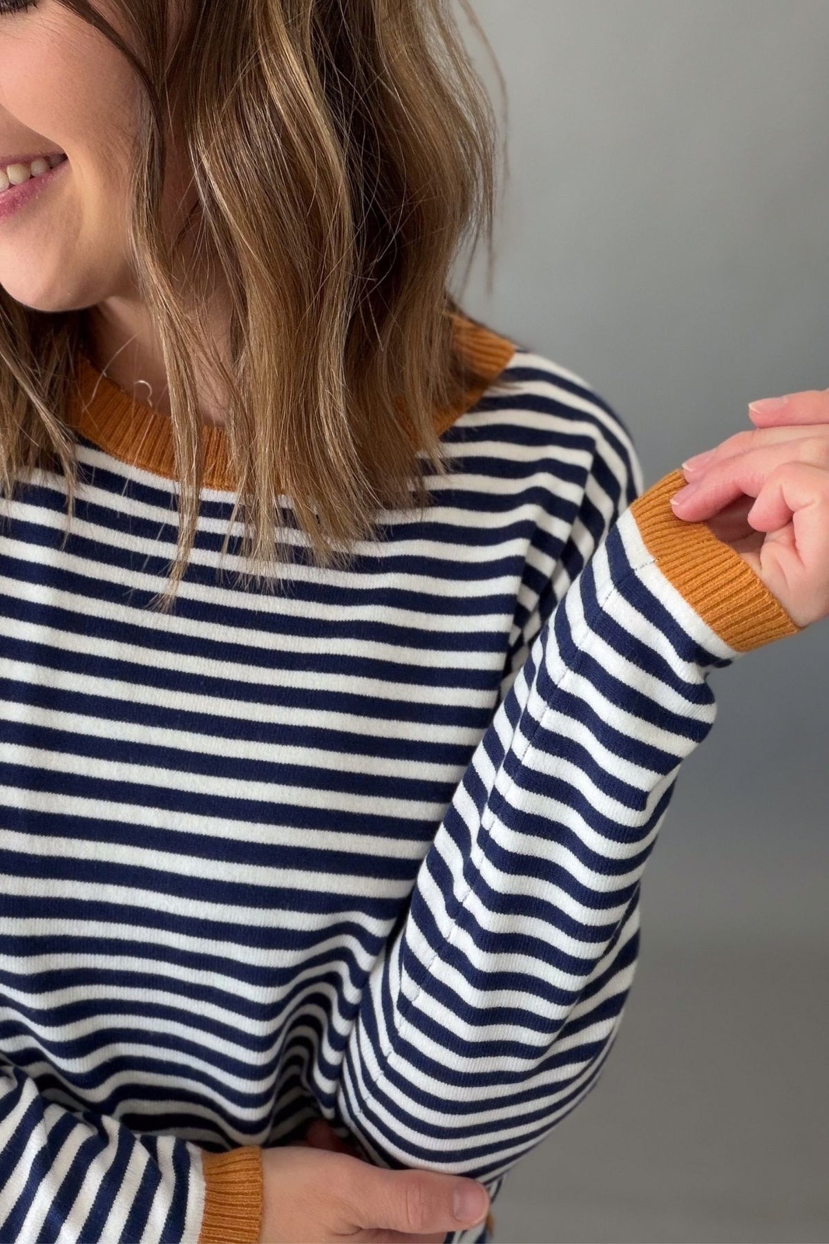 A close-up of a woman wearing a navy and white horizontally striped knit sweater with contrasting mustard yellow ribbed trim at the crew neck and cuffs, against a plain gray background.
