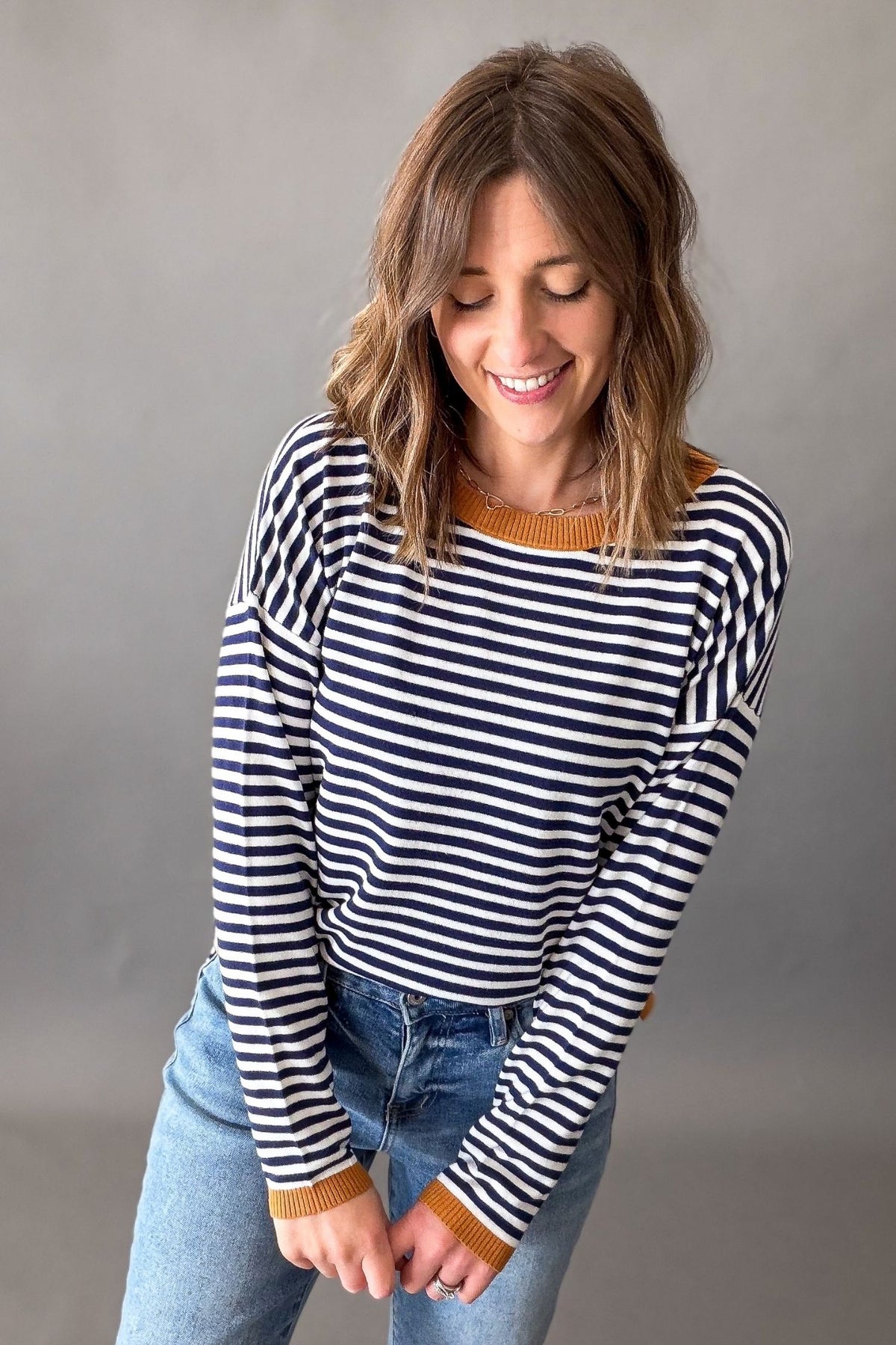 A woman wearing a navy and white horizontally striped long-sleeve knit sweater with a mustard yellow crew neck and cuffs, paired with blue jeans, posing against a neutral gray studio background.
