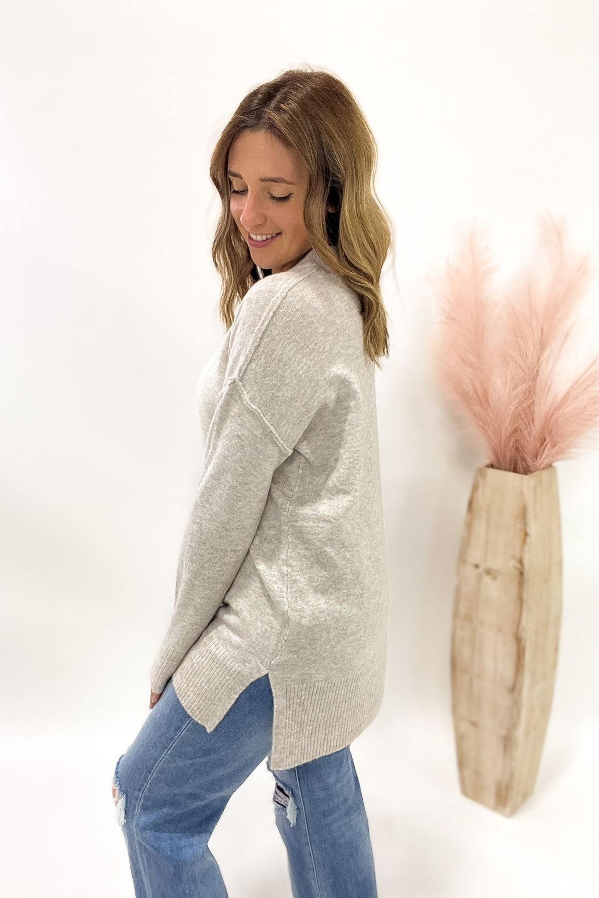 Woman wearing a oatmeal sweater and blue jeans standing next to a decorative plant against a white background