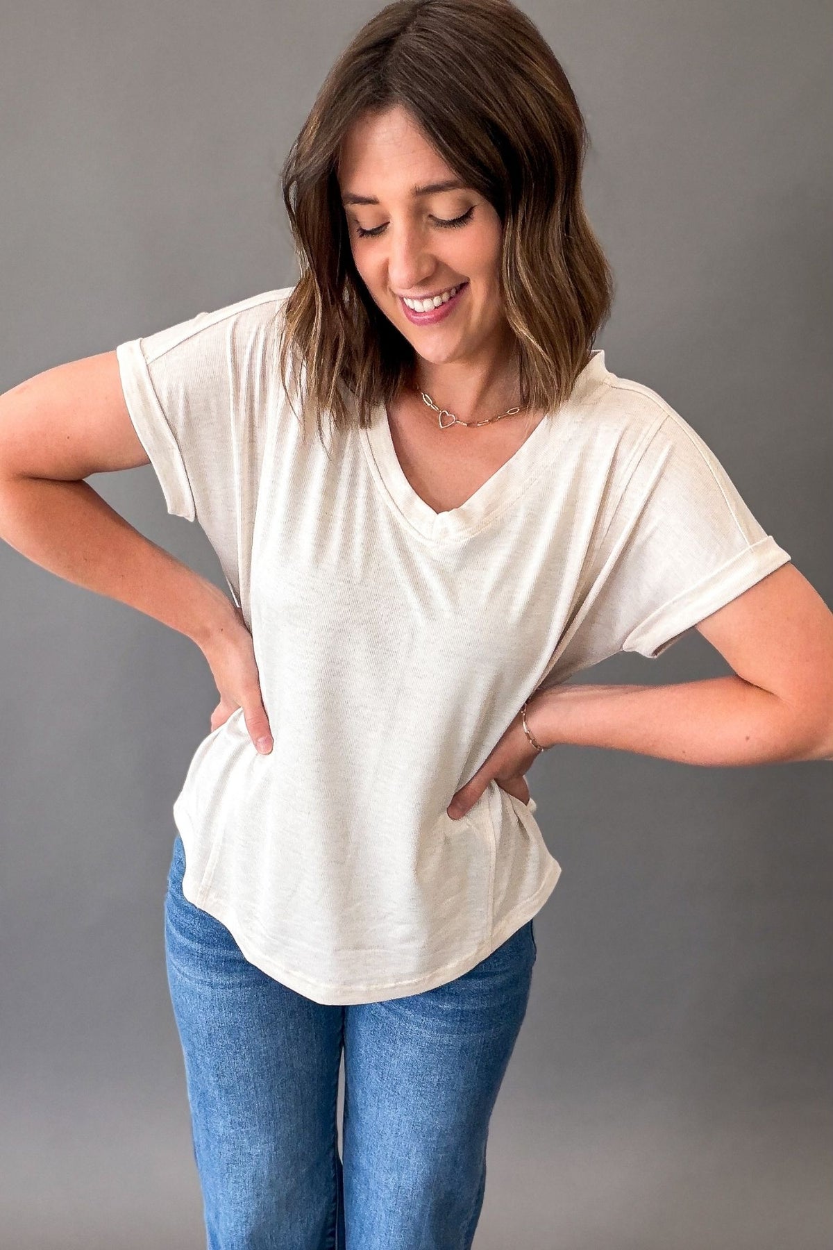 Woman wearing a oatmeal t-shirt and blue jeans against a gray background