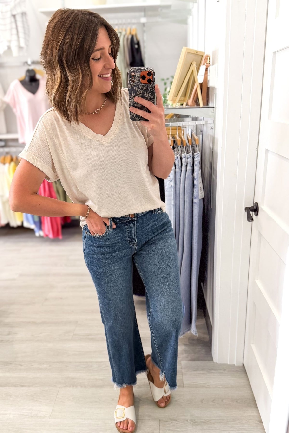 Woman taking a mirror selfie wearing a oatmeal t-shirt and blue jeans in a clothing store.