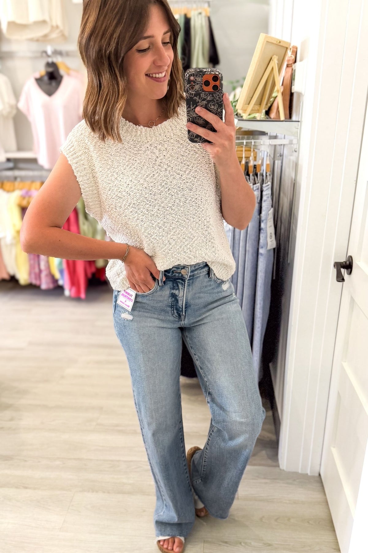 woman taking a selfie in a boutique wearing an off white textured short sleeve knit sweater and blue jeans