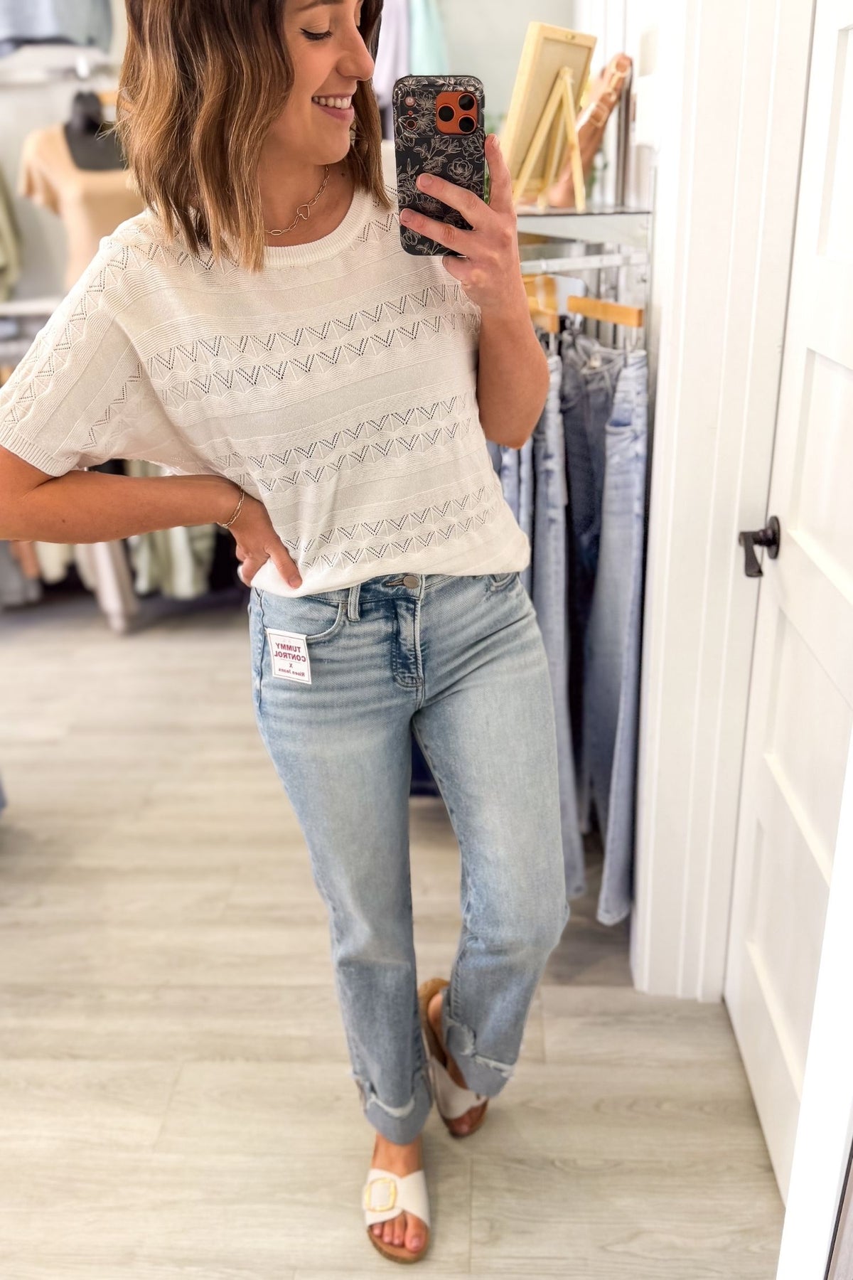 Woman taking a mirror selfie wearing a white pointelle knit top and light blue jeans in a clothing store.