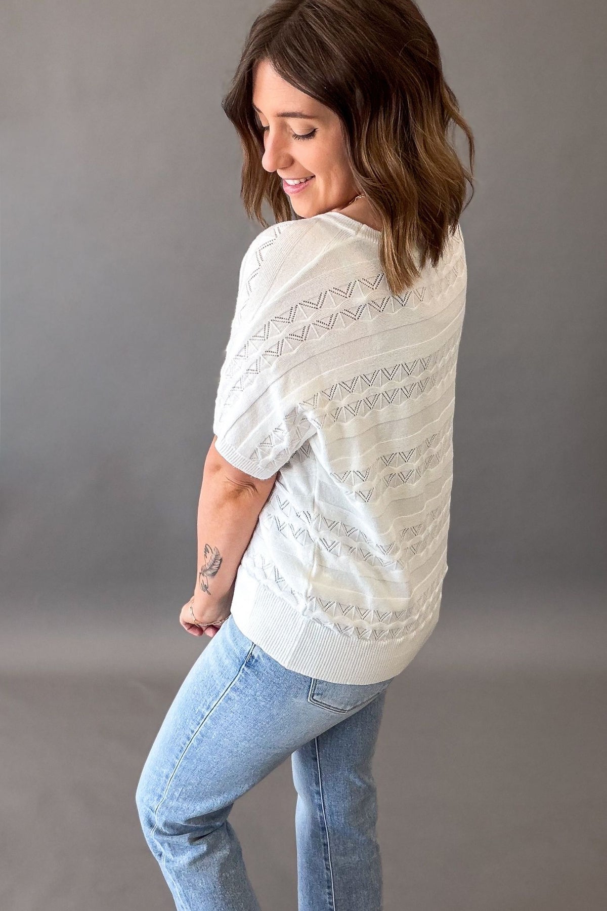 Woman wearing an off white pointelle knit short sleeve top with lightweight soft fabric and stretch, casual versatile women’s top showing back details.