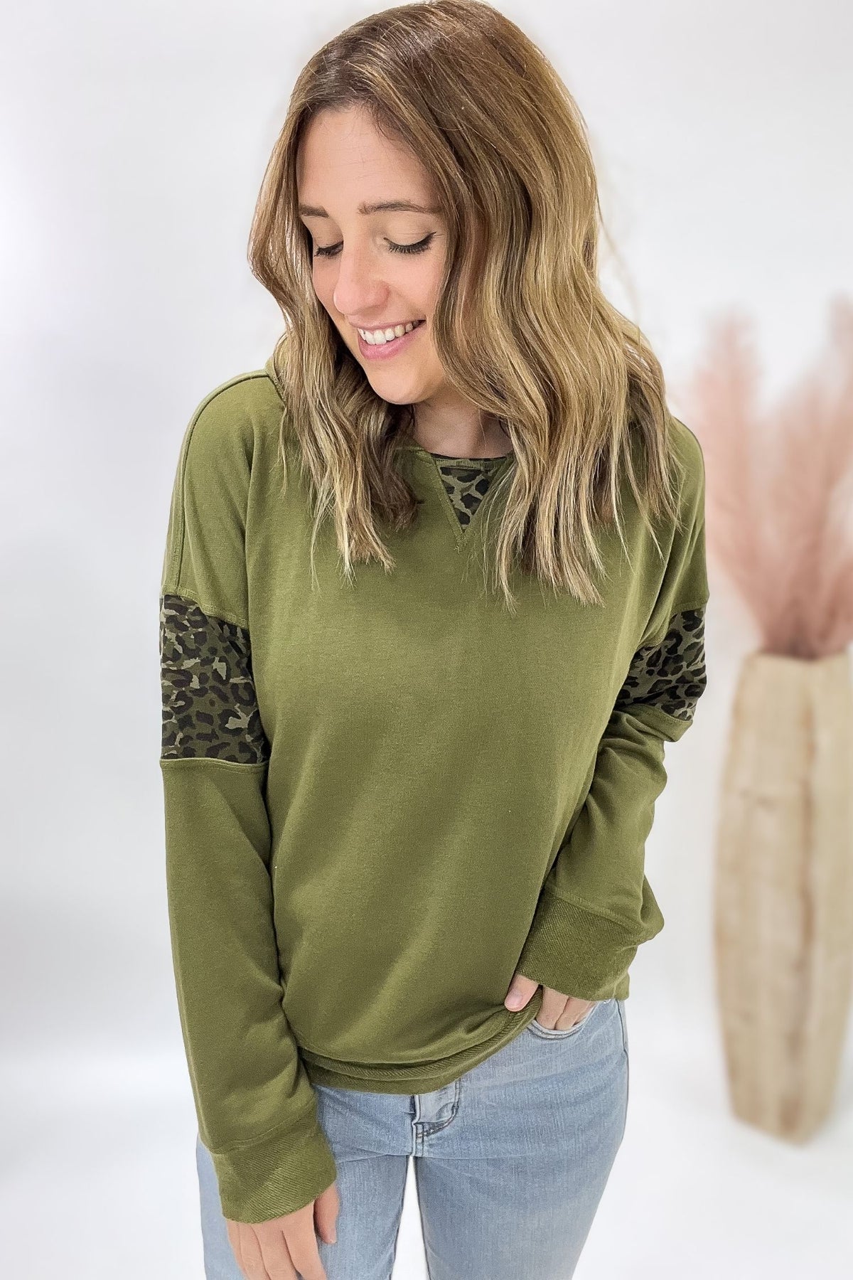 Woman wearing an olive green long-sleeve top with leopard print sleeves on a white background