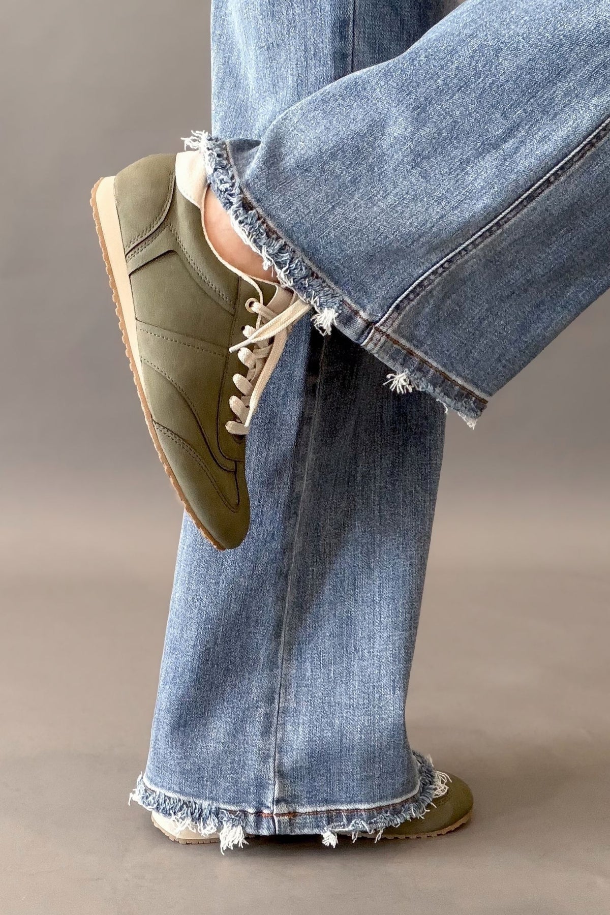An olive green sneaker with cream laces and a beige sole, worn by a model in light-wash frayed-hem jeans against a neutral studio background.