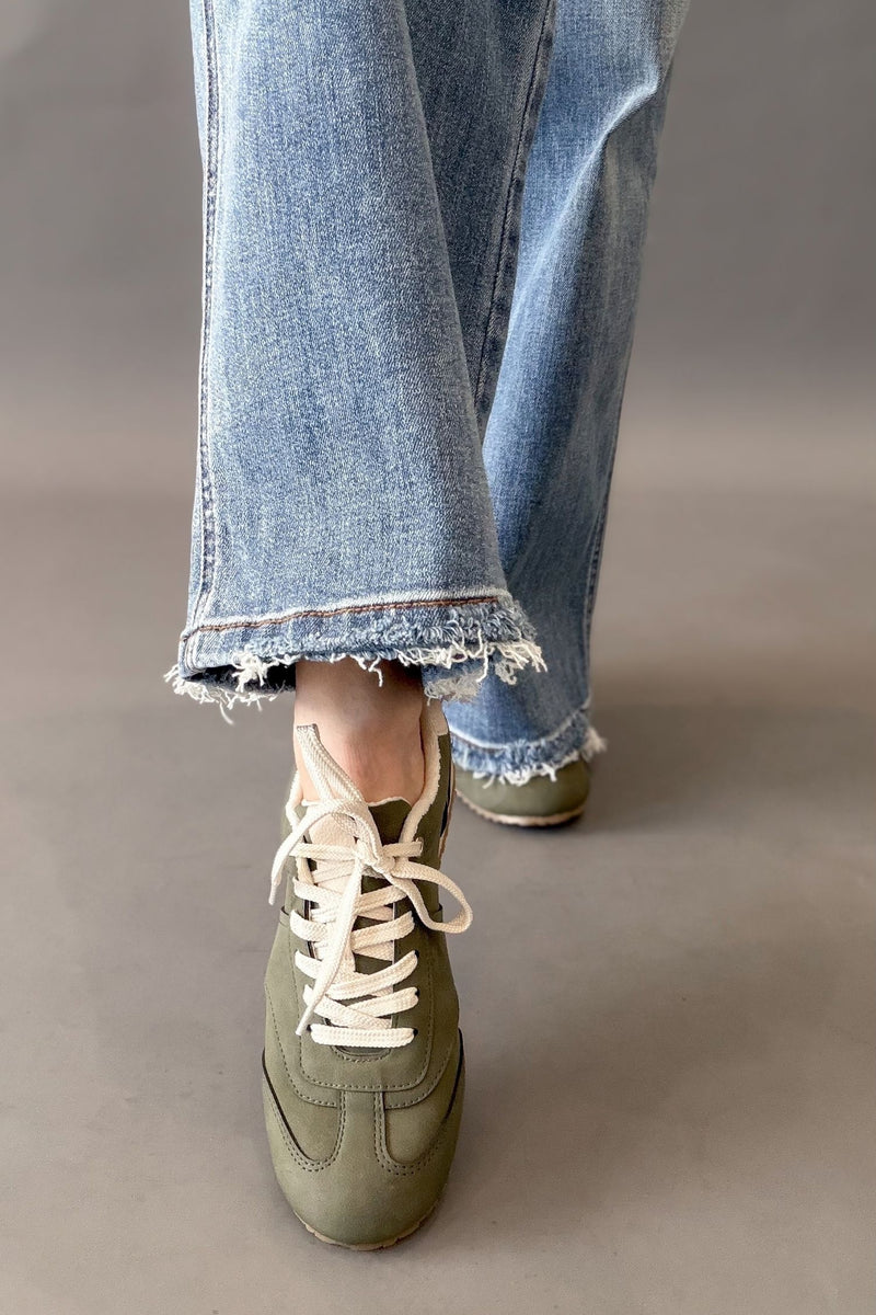A person wearing light blue frayed-hem jeans and olive green low-top sneakers with white laces, shown from the calves down against a plain gray studio background.