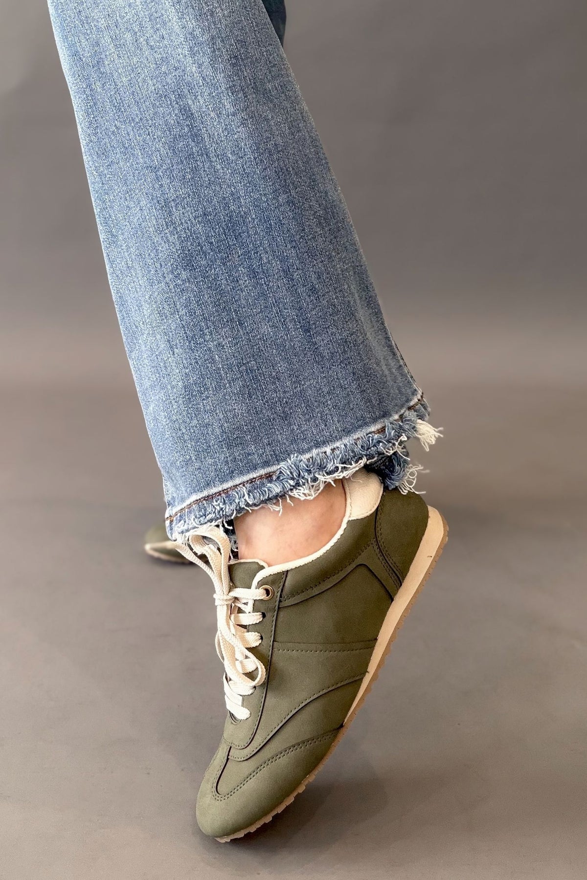 An olive green sneaker with white laces and a beige sole, worn with light-wash frayed-hem jeans against a neutral gray background.