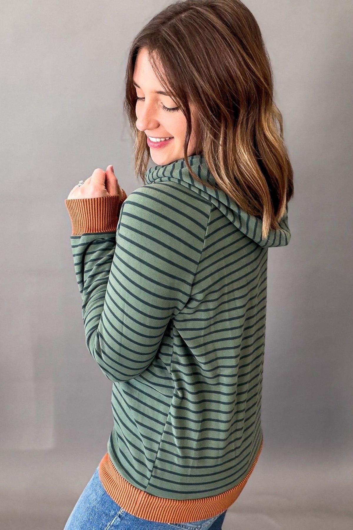 A woman wearing a olive green and dark green striped hoodie with contrasting brown ribbed cuffs, hem, and collar, shown from a three-quarters back view against a neutral gray studio background.