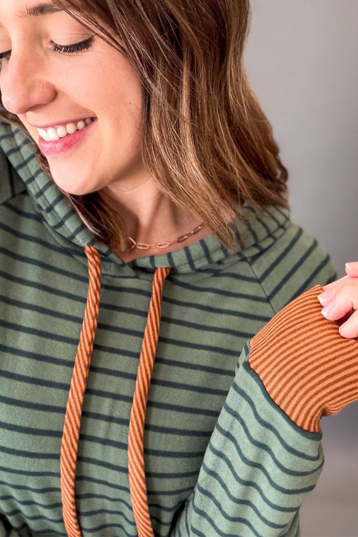 A close-up lifestyle shot of a woman wearing a olive green and black striped hoodie with orange rope drawstrings and matching orange ribbed elbow patches.
