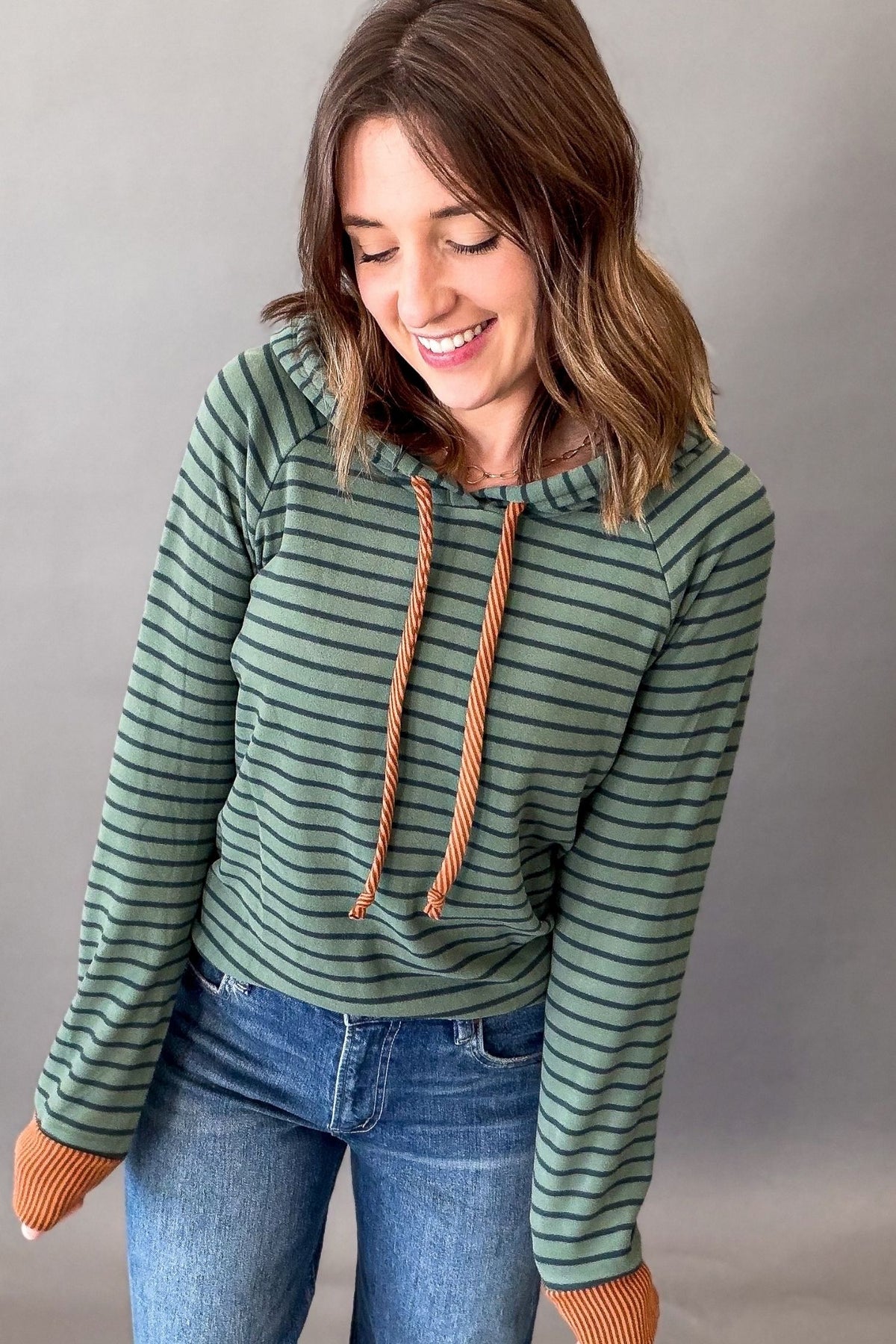 A woman wearing a green and dark green striped long-sleeve hoodie with orange drawstrings and matching orange ribbed cuffs, paired with blue jeans, against a neutral gray background.