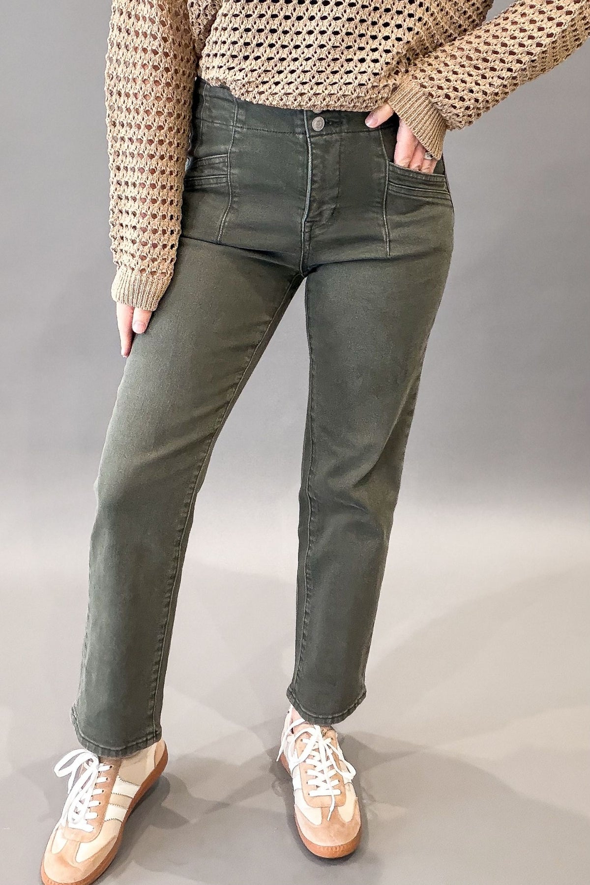 A person wearing olive green straight-leg jeans, a beige knit sweater, and beige sneakers, standing against a neutral gray studio background.