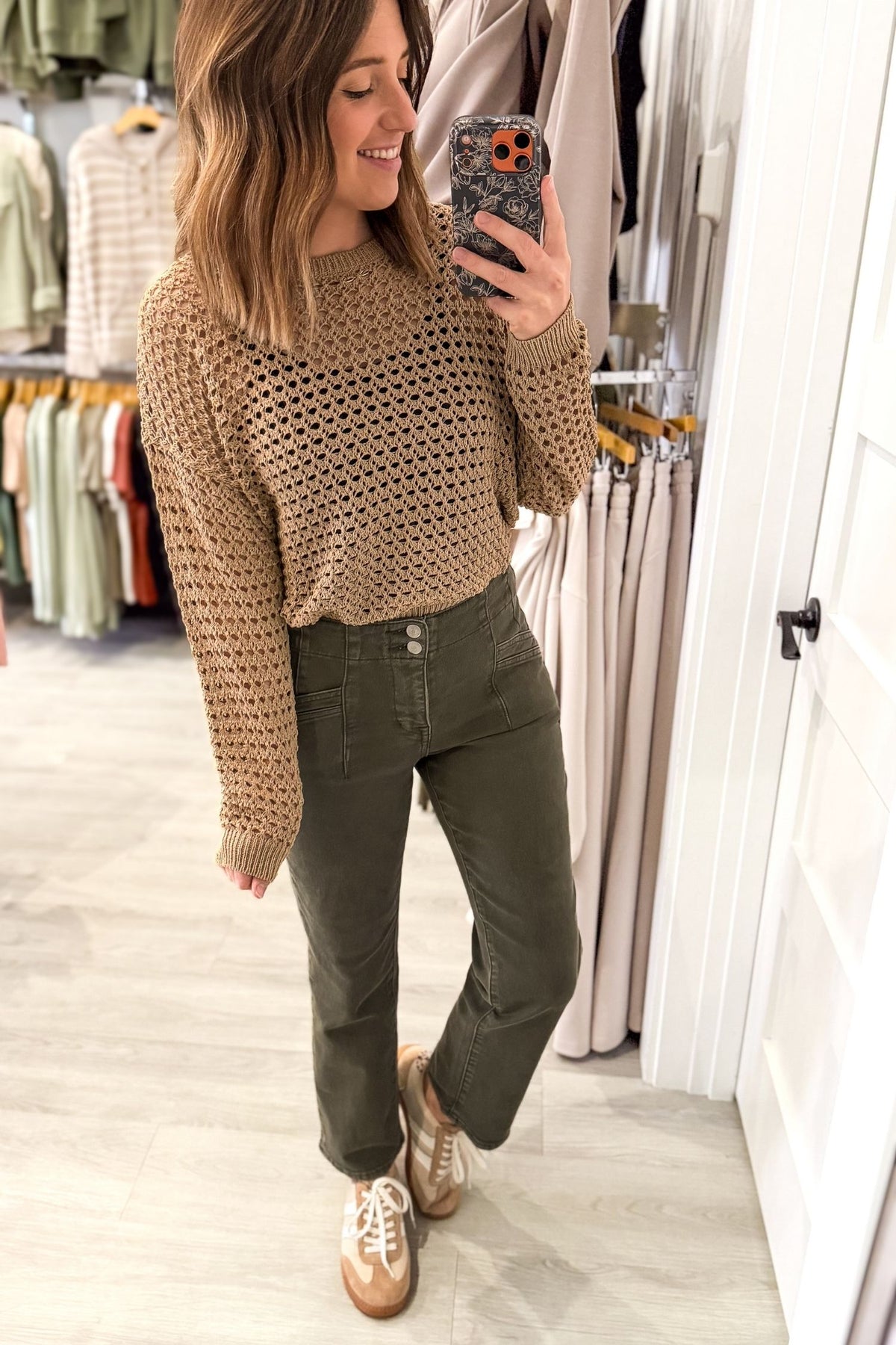 A woman takes a mirror selfie in a clothing boutique, wearing a beige open-knit long-sleeve sweater and olive green pants with brown sneakers.