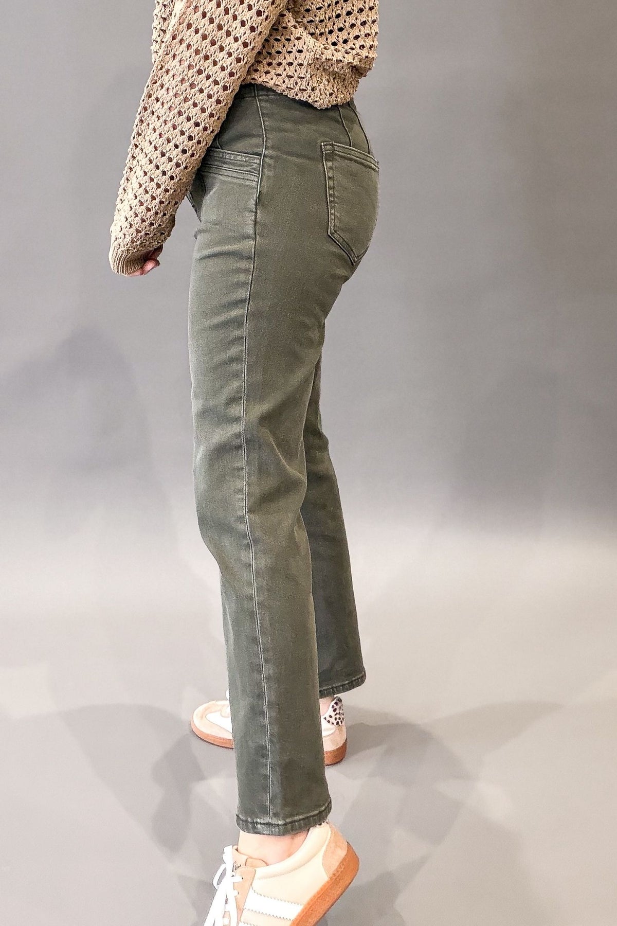 A person shown from the back wearing olive green straight-leg jeans, a beige crochet knit top, and white sneakers with orange soles, standing against a gray studio background.