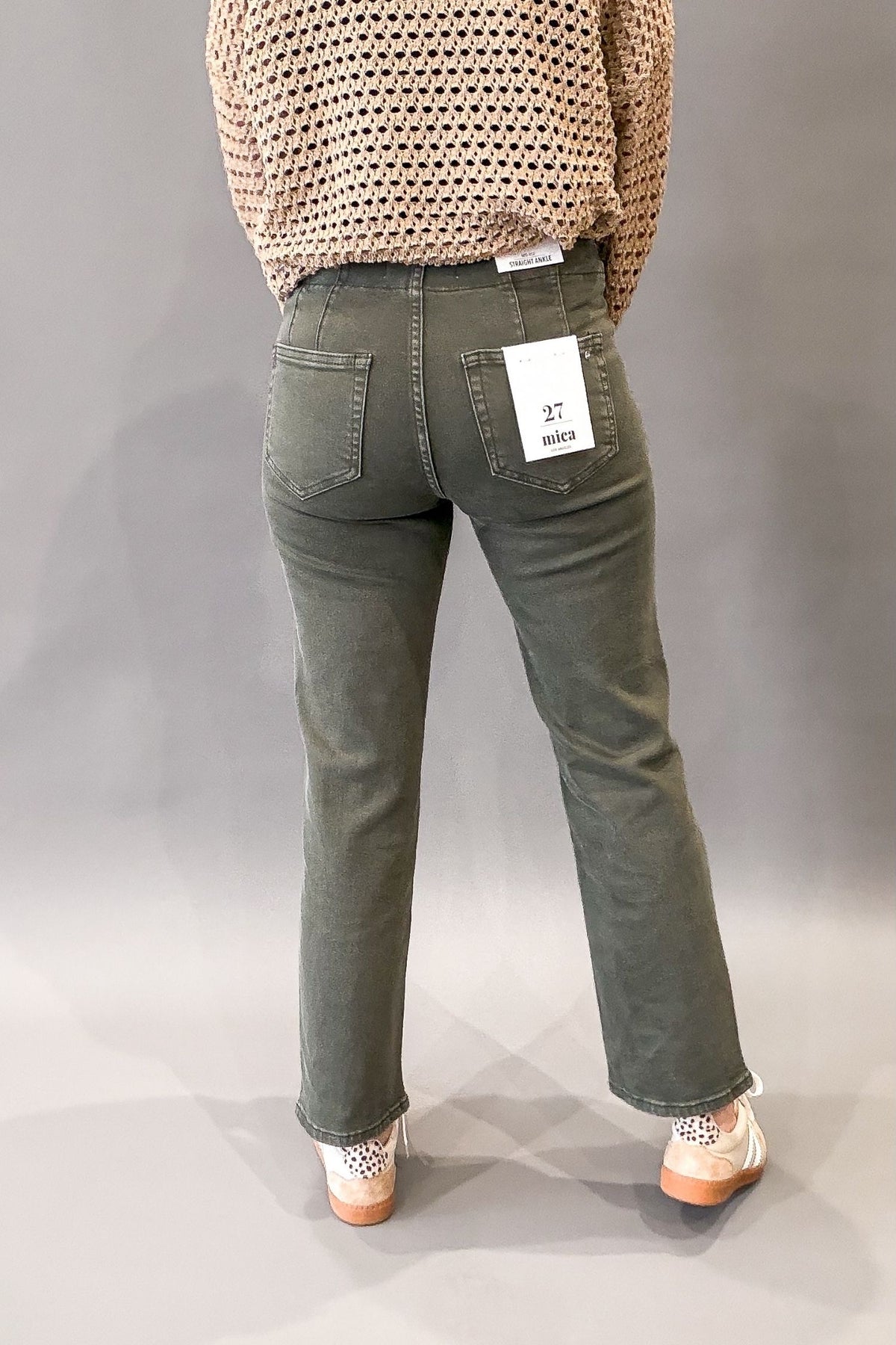 A person shown from the back wearing olive green straight-leg jeans, a beige crochet knit top, and white sneakers with orange soles, standing against a neutral gray studio background.