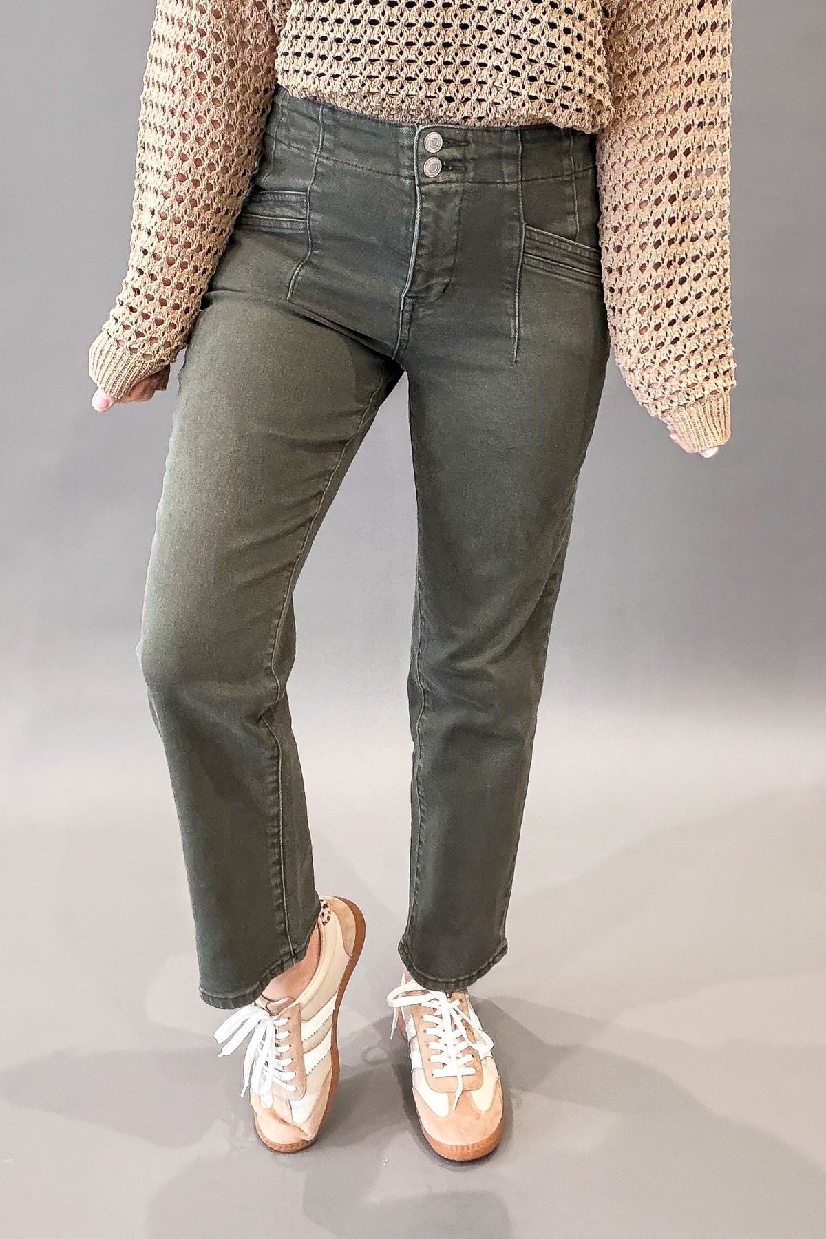 A person wearing olive green straight-leg jeans, a beige chunky knit sweater, and beige sneakers, shown from the chest down against a neutral gray studio background.
