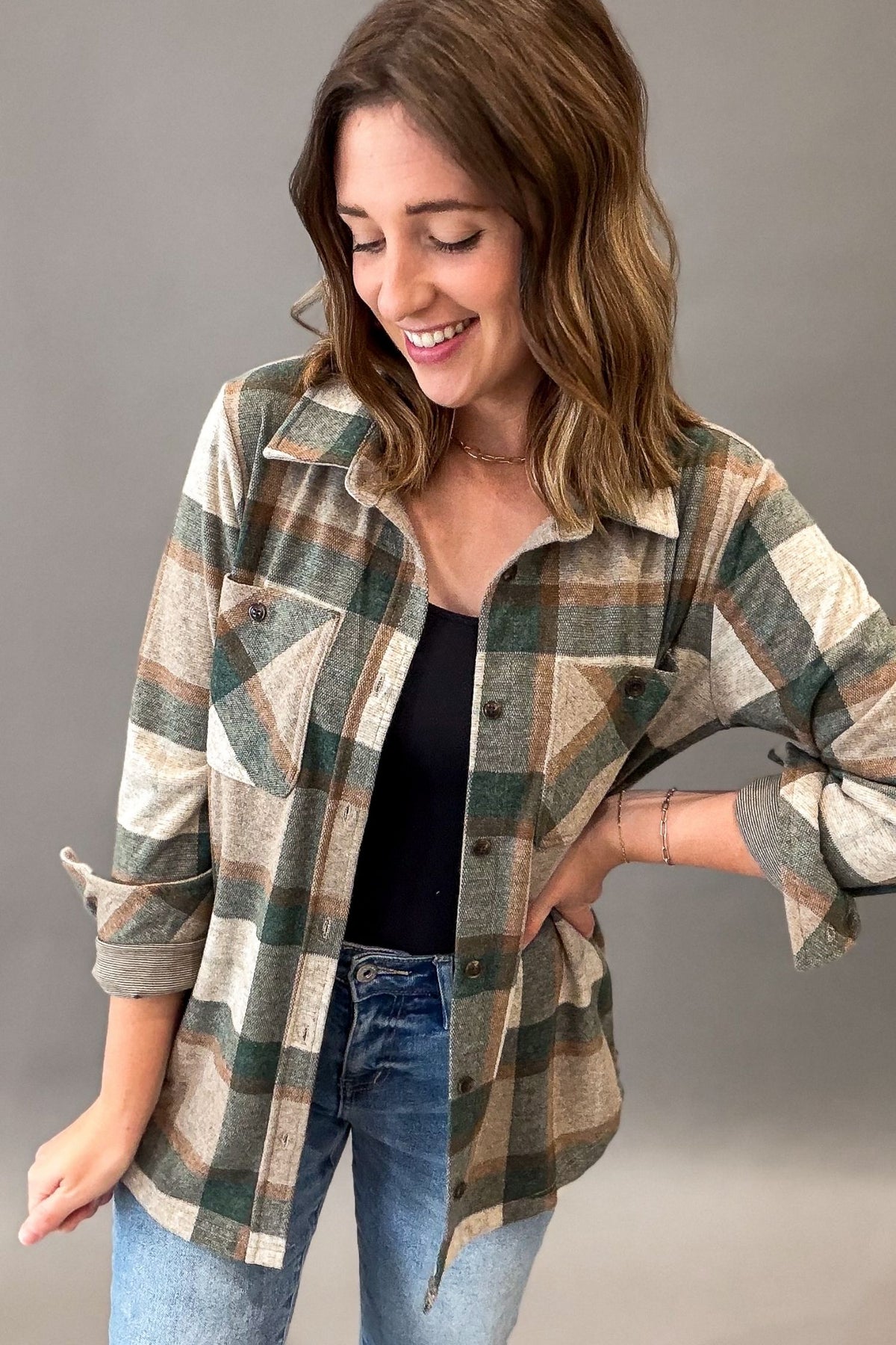 A woman wearing a long-sleeve plaid button-up shirt in green, beige, and brown tones, layered over a black top and blue jeans, photographed against a plain gray studio background.