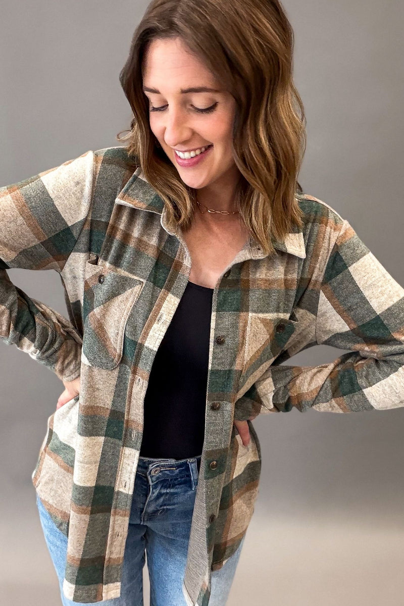 A woman wearing a long-sleeve plaid button-up flannel shirt in green, beige, and brown tones, layered over a black top and blue jeans, posing against a neutral gray studio background.