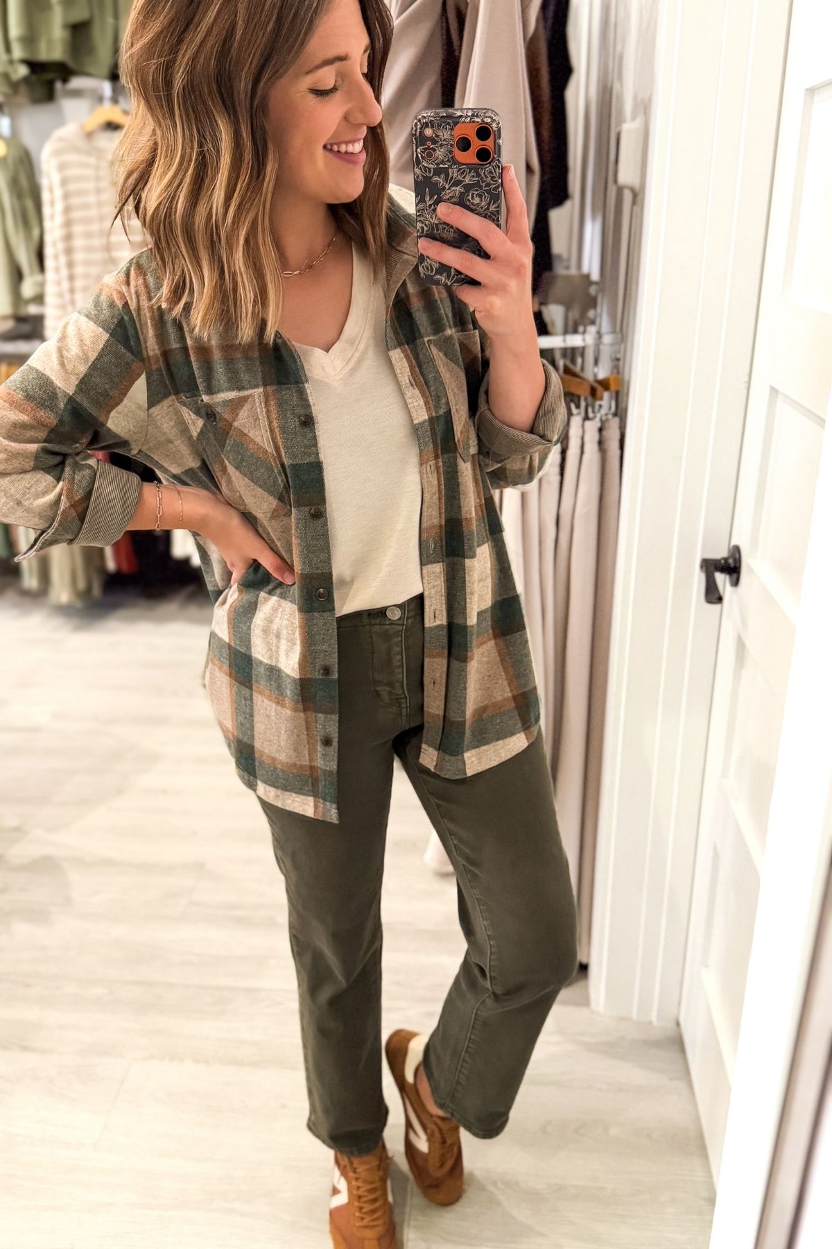 A woman takes a mirror selfie in a boutique while wearing a green and beige plaid button-up shirt over a cream top, olive green pants, and brown lace-up boots.