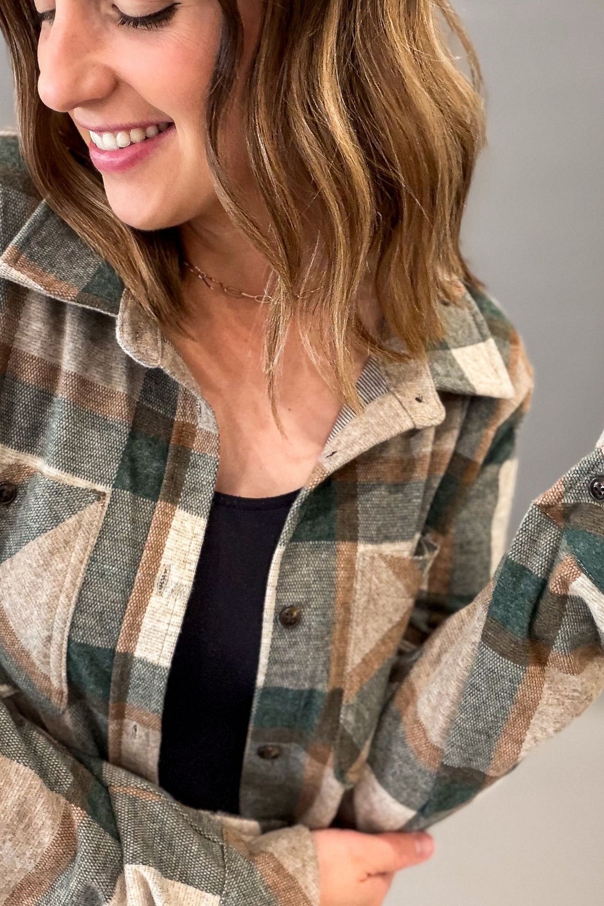 A woman wearing a plaid button-up flannel shirt in muted green, beige, and brown tones over a black top, shown in a close-up lifestyle shot against a plain gray background.