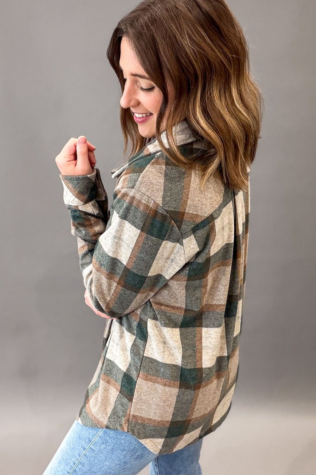 A woman wearing a long-sleeve plaid flannel shirt in beige, green, and brown, paired with light blue jeans, photographed against a neutral gray studio background.