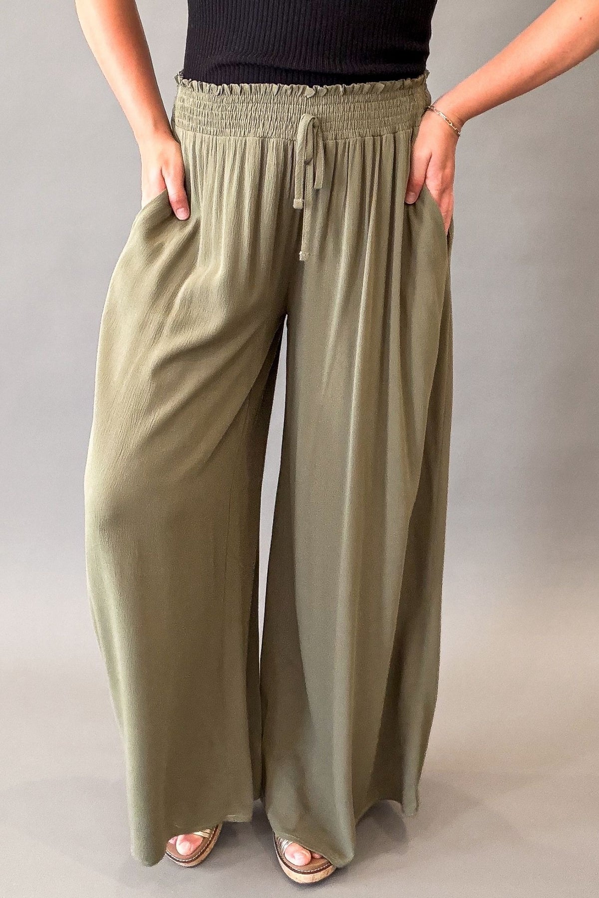 A person wearing loose, wide-leg sage green pants with an elastic drawstring waistband, standing against a plain gray studio background.