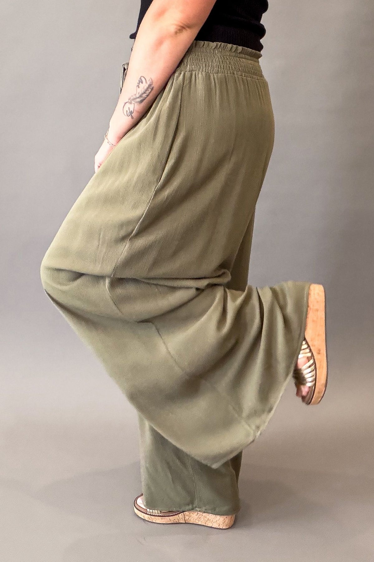 woman wearing olive smocked waistband wide leg pants and wedge sandals with a black tank showing the side and back details