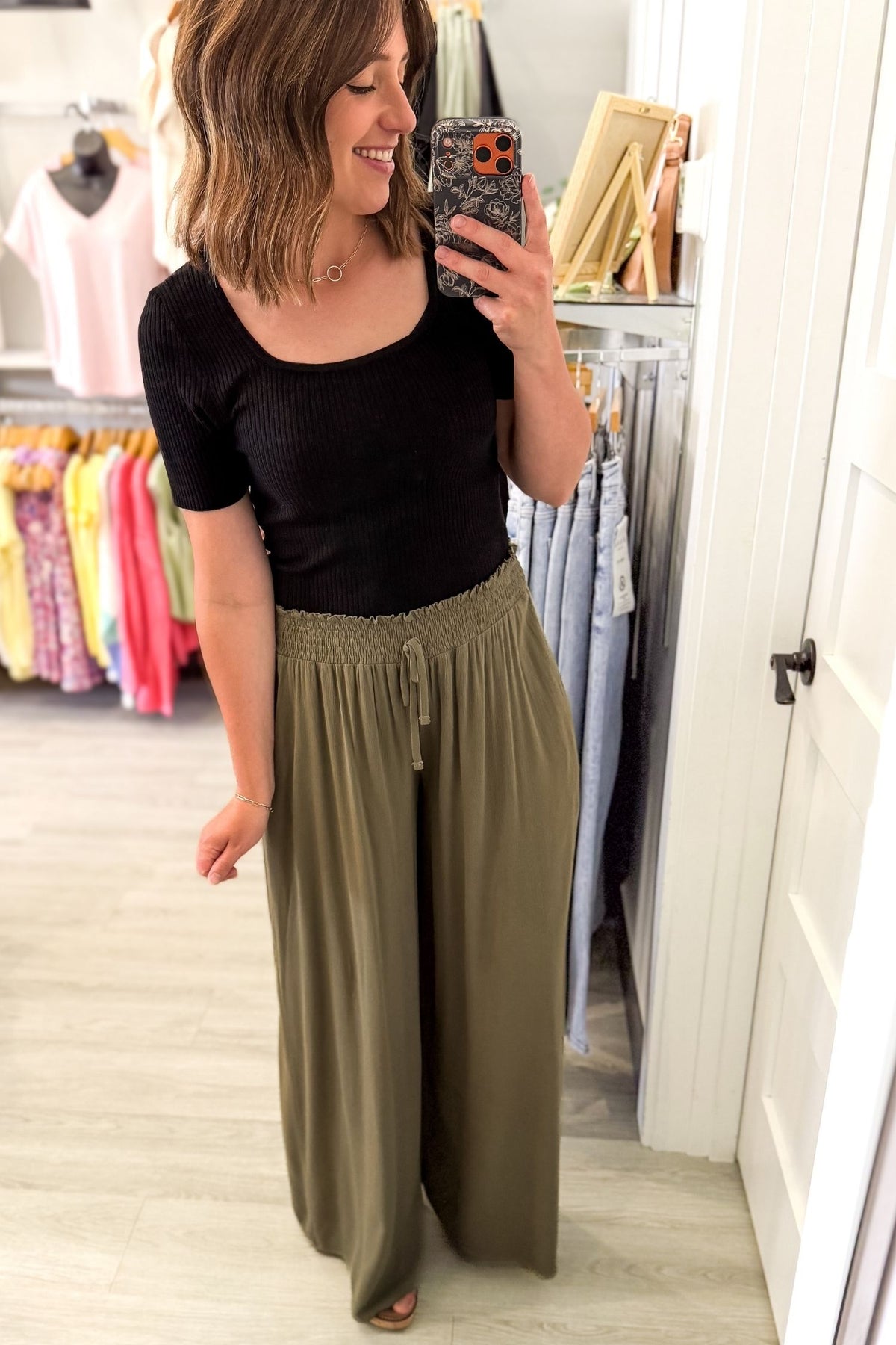 woman taking  selfie in a boutique wearing olive smocked waistband wide leg pants and wedge sandals with a black tank