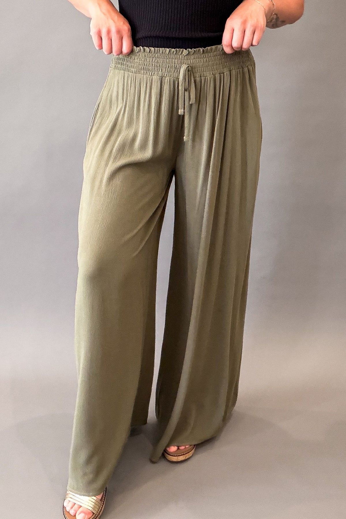 woman wearing olive smocked waistband wide leg pants and wedge sandals with a black tank