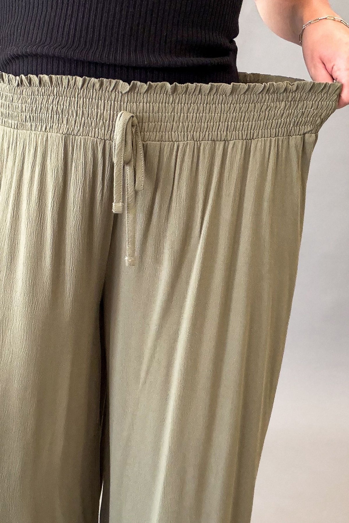 Close-up of a person wearing loose, wide-leg faux drawstring pants in a olive color, with the hand stretching the elastic waistband.