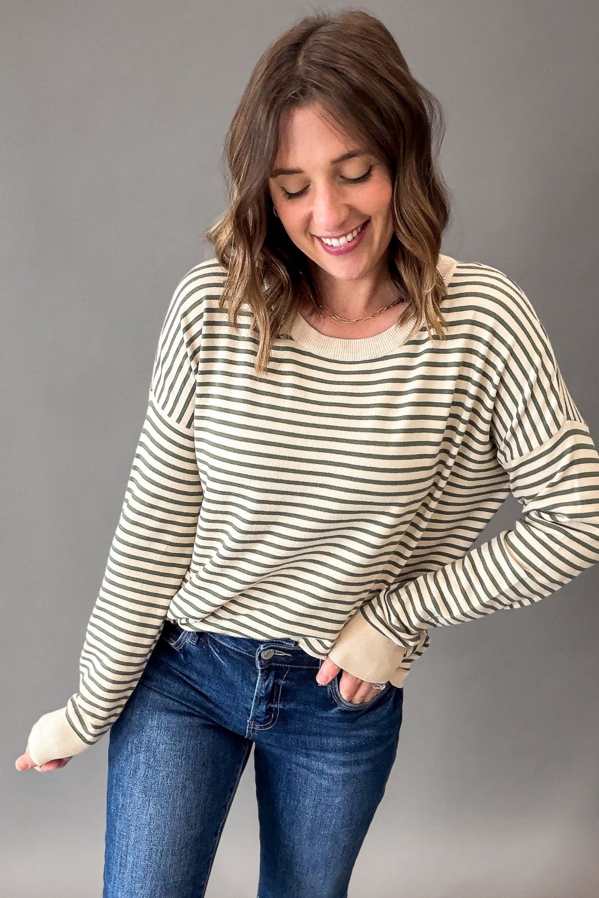 A woman wearing a cream and dark brown horizontally striped long-sleeve knit top with ribbed cuffs, paired with blue jeans, standing against a solid gray studio background.