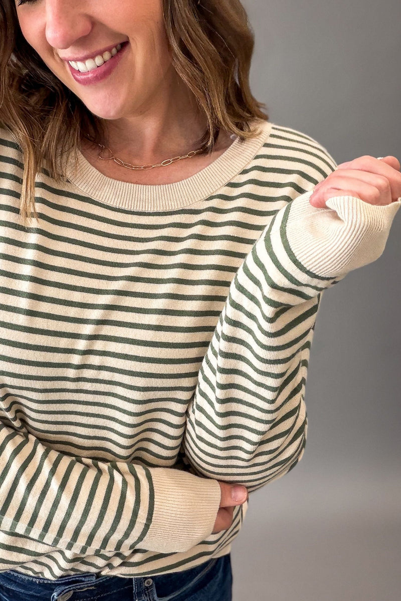 A woman wearing a cream and dark green striped long-sleeve knit sweater, shown from the chest down against a plain gray studio background.