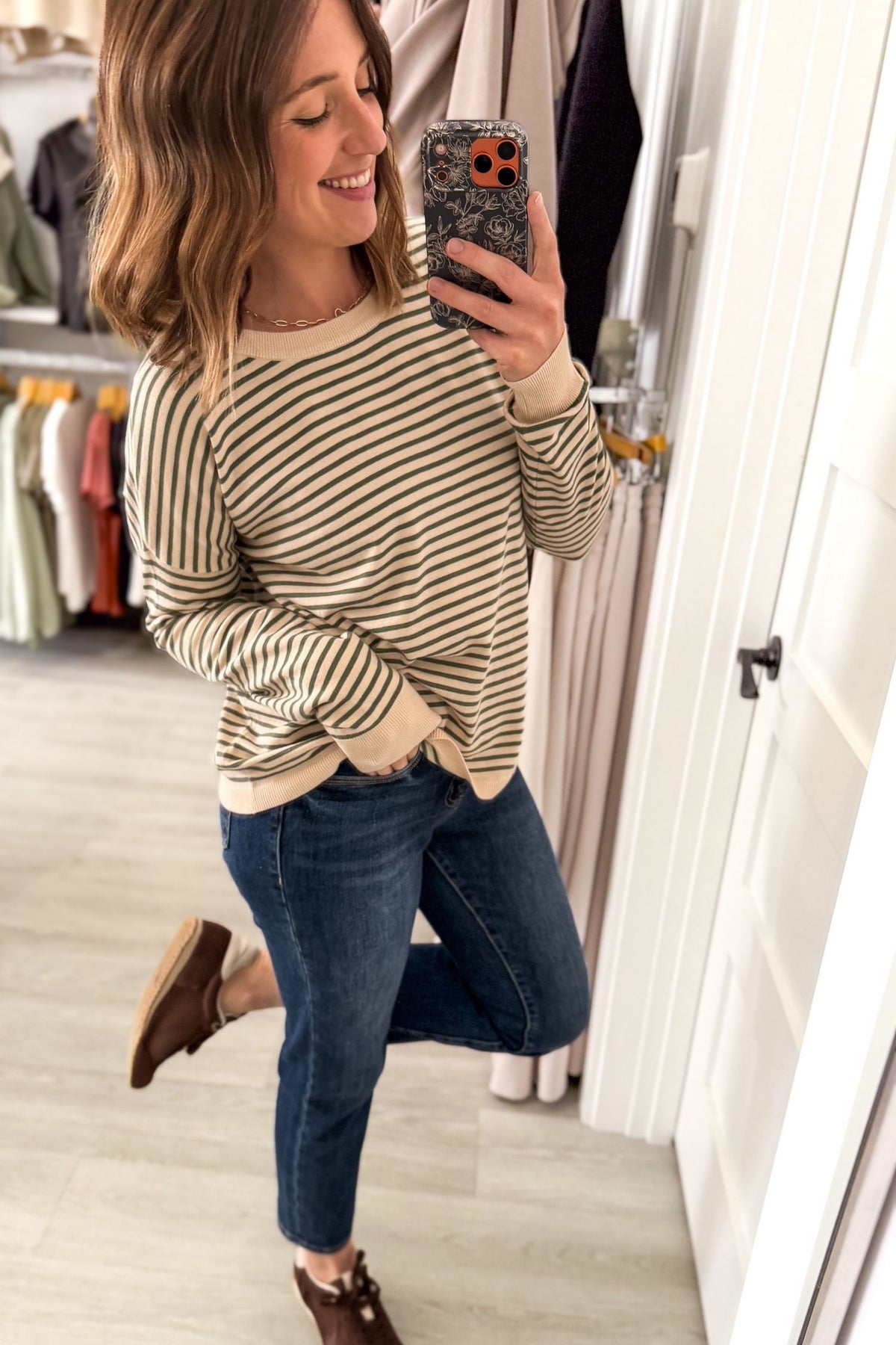 A woman takes a mirror selfie in a clothing store, wearing a beige and dark brown horizontally striped long-sleeve knit sweater, blue jeans, and brown lace-up shoes.