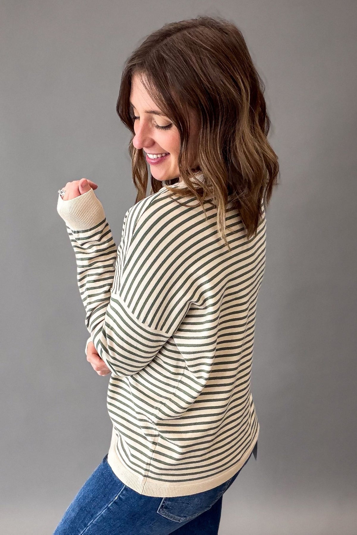 A woman wearing a cream and dark brown horizontally striped long-sleeve knit top, paired with blue jeans, photographed against a solid gray studio background.