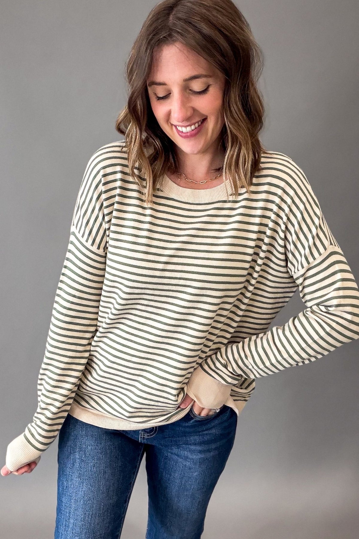 A woman wearing a cream and dark brown horizontal striped long-sleeve crewneck sweater, paired with blue jeans, standing against a solid gray studio background.