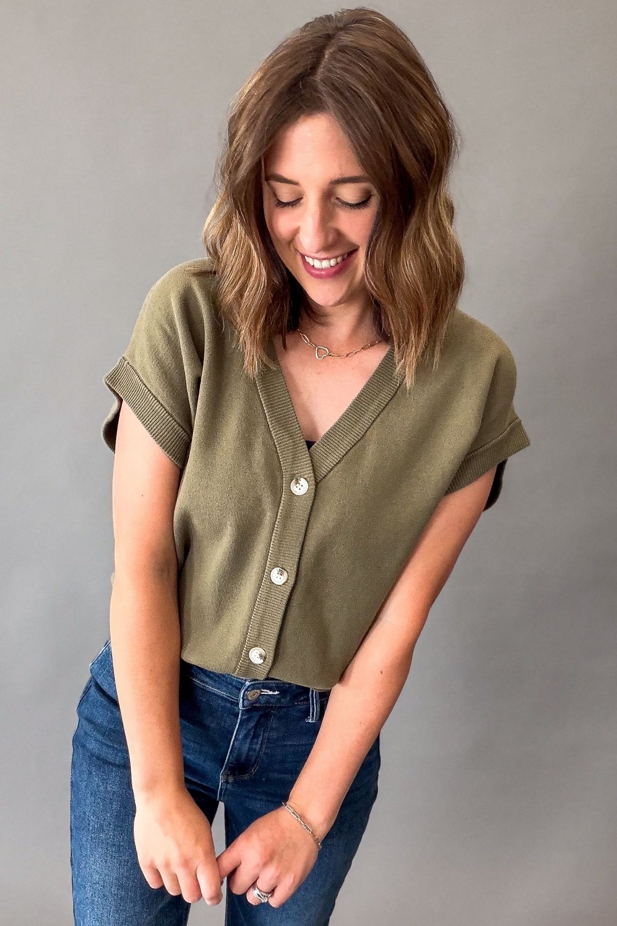 A woman wearing a short-sleeve olive green button-up knit top and blue jeans, standing against a plain gray studio background.
