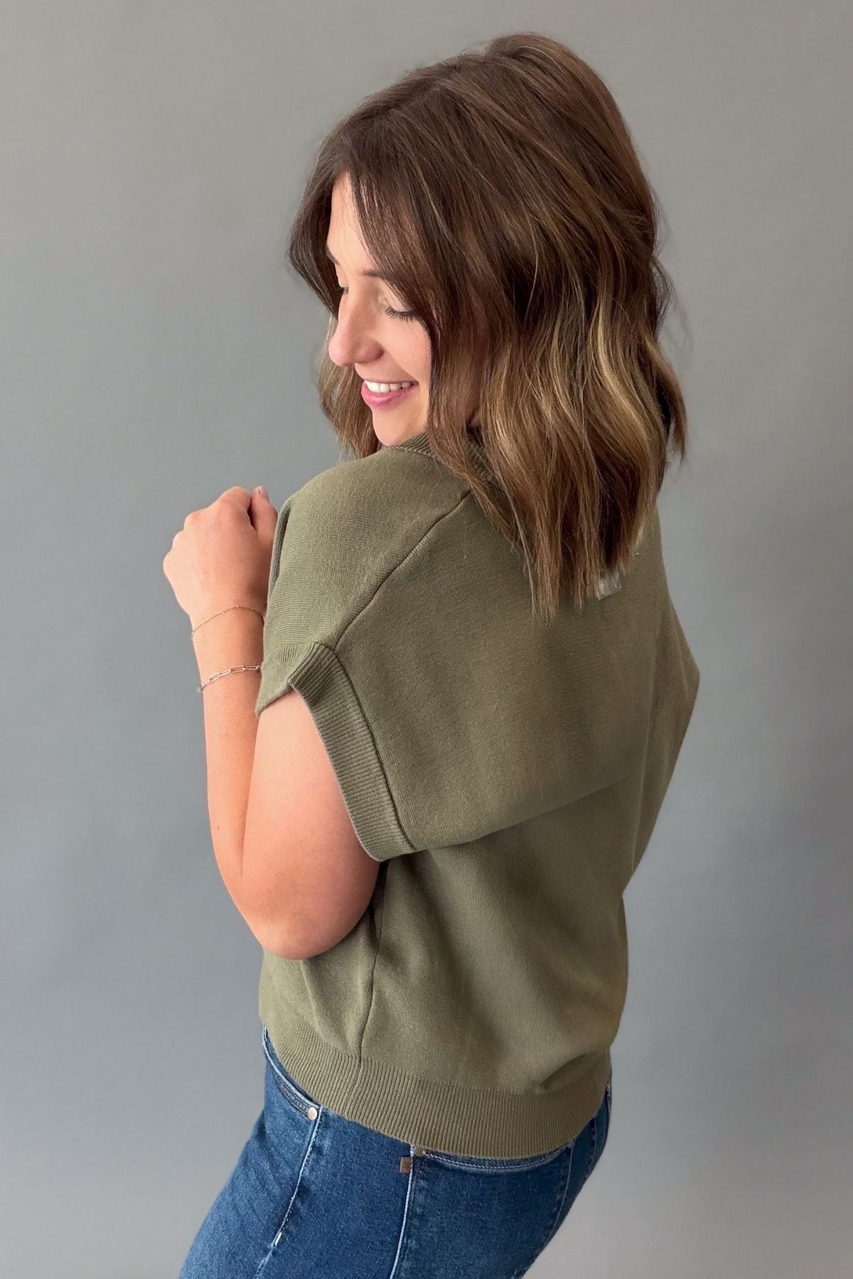 A woman with shoulder-length brown hair is shown from the back wearing an olive green short-sleeve knit top and blue jeans against a plain gray studio background.
