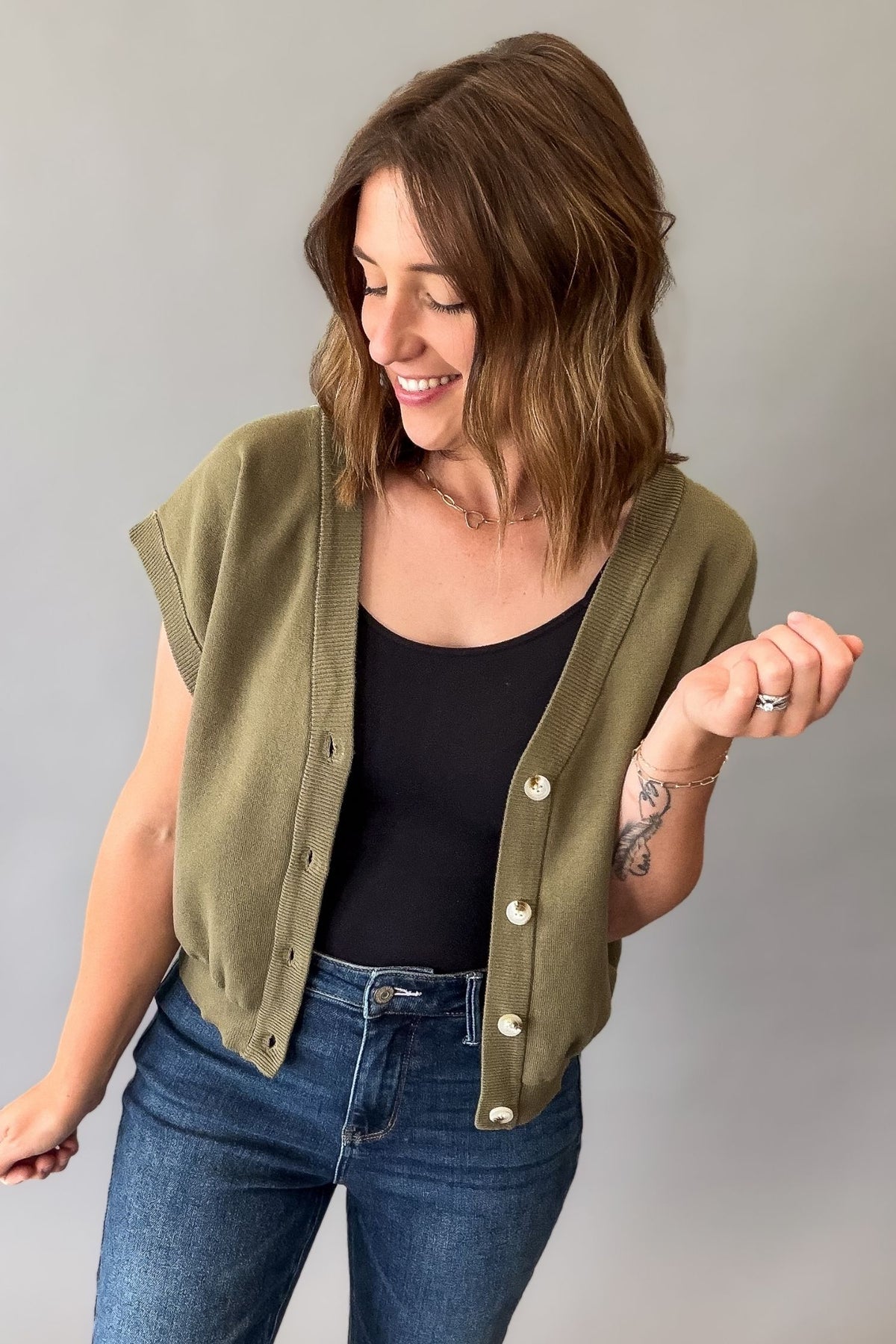 A woman wearing an olive green sleeveless button-front cardigan over a black tank top and blue jeans, standing against a plain light gray studio background.