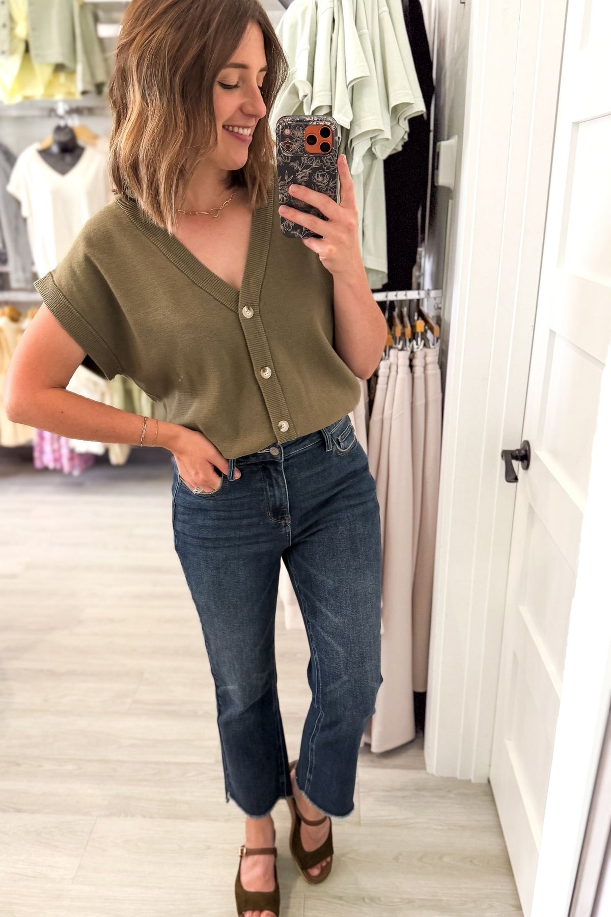A woman takes a mirror selfie in a boutique wearing an olive green short-sleeve button-up top and dark blue cropped jeans.