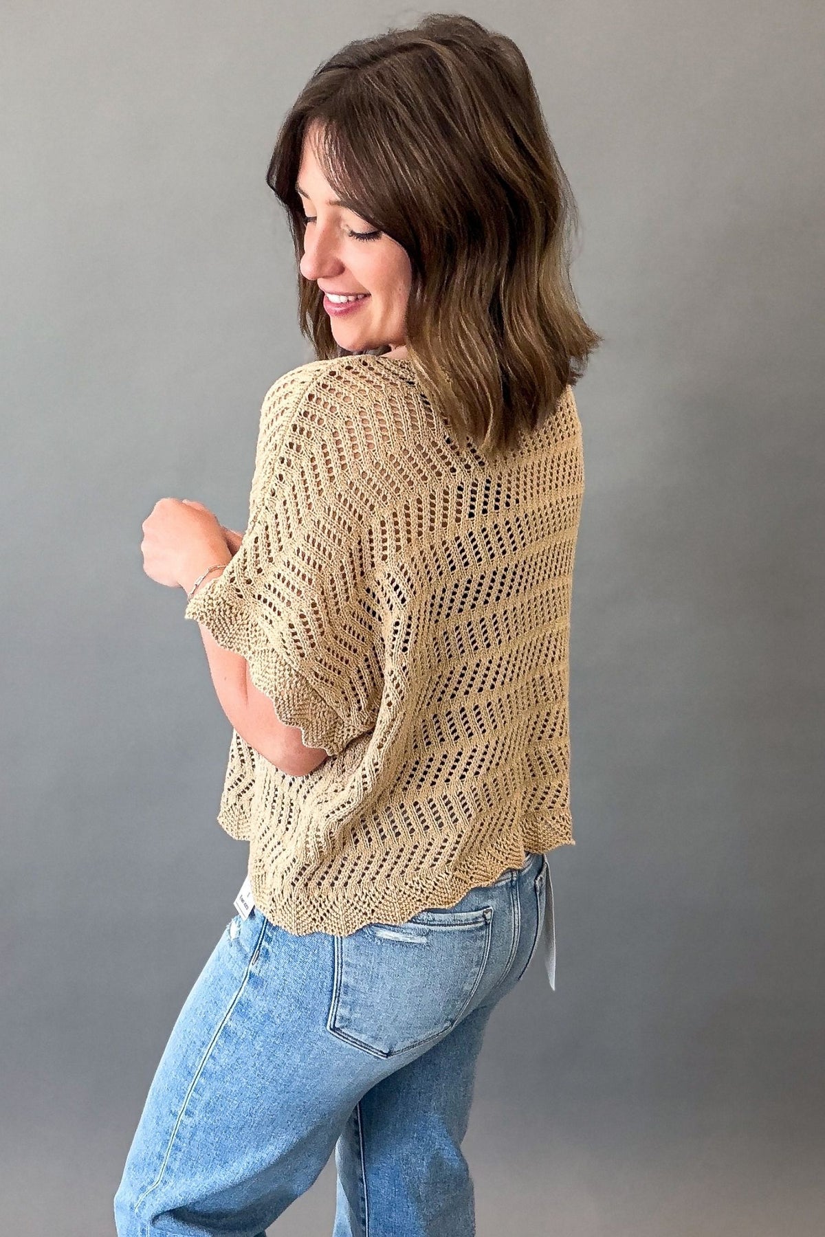 Woman wearing a lightweight latte open weave crochet sweater with short sleeves styled with light wash denim jeans showing the back and side details.