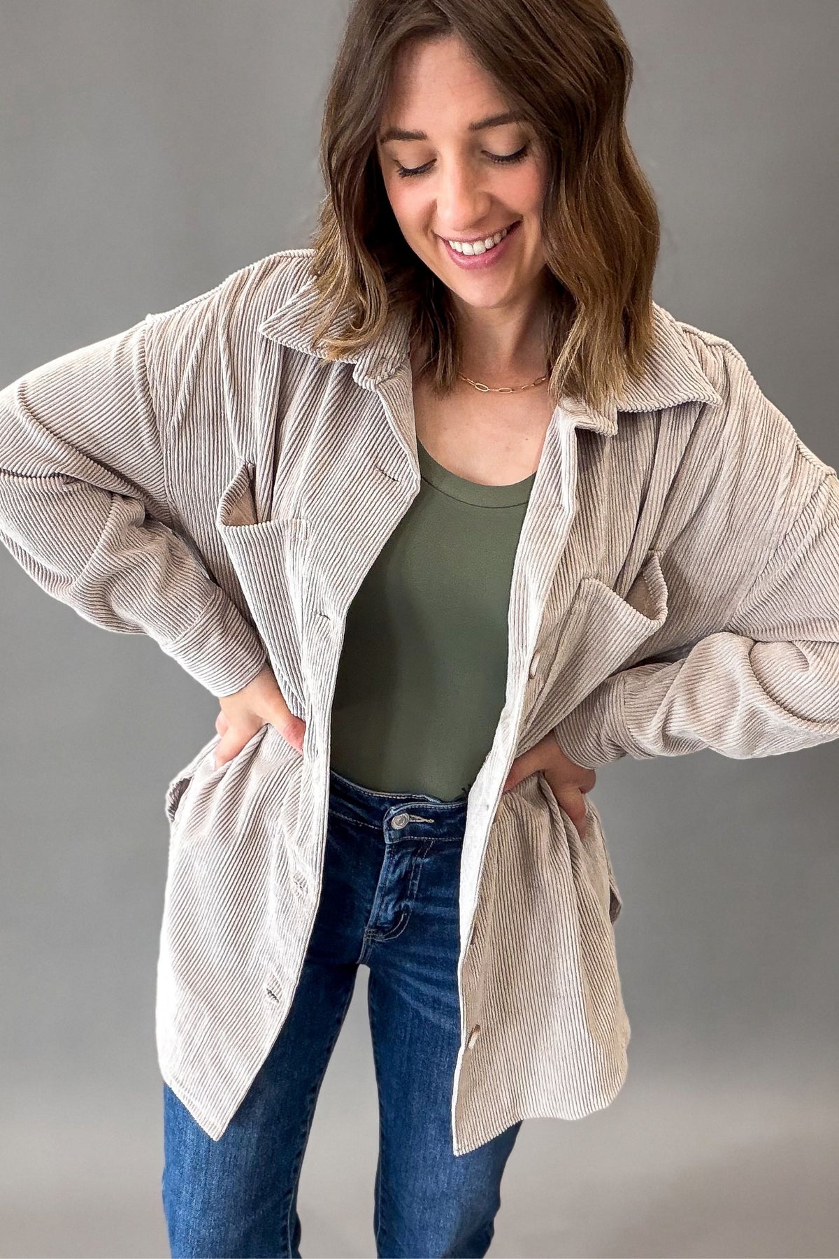 A woman wearing a light beige corduroy button-up shirt jacket over an olive green tank top and blue jeans, posing against a neutral gray studio background.