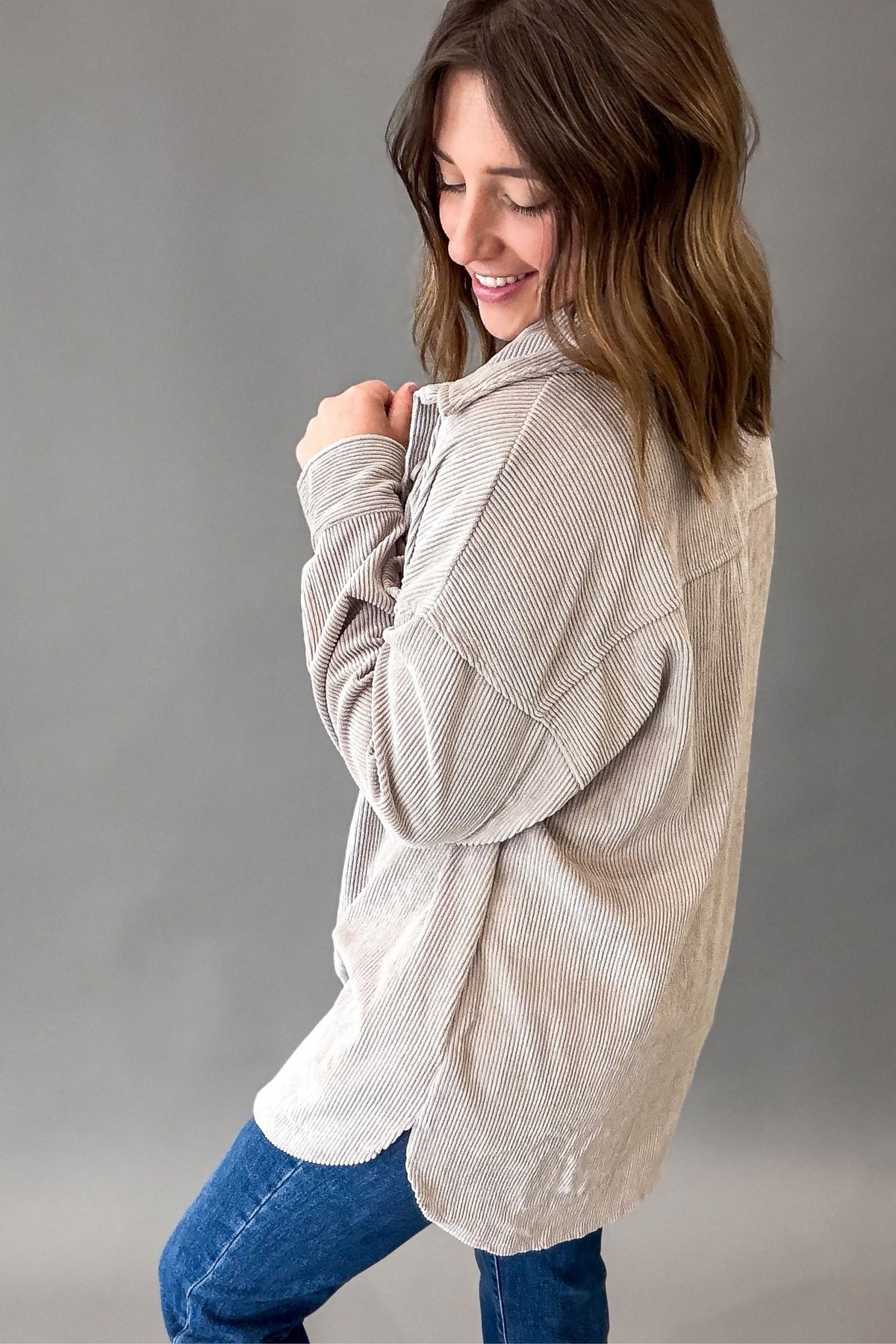 A woman wearing a light beige corduroy button-up shirt with blue jeans, shown from a side-back angle against a neutral gray studio background.