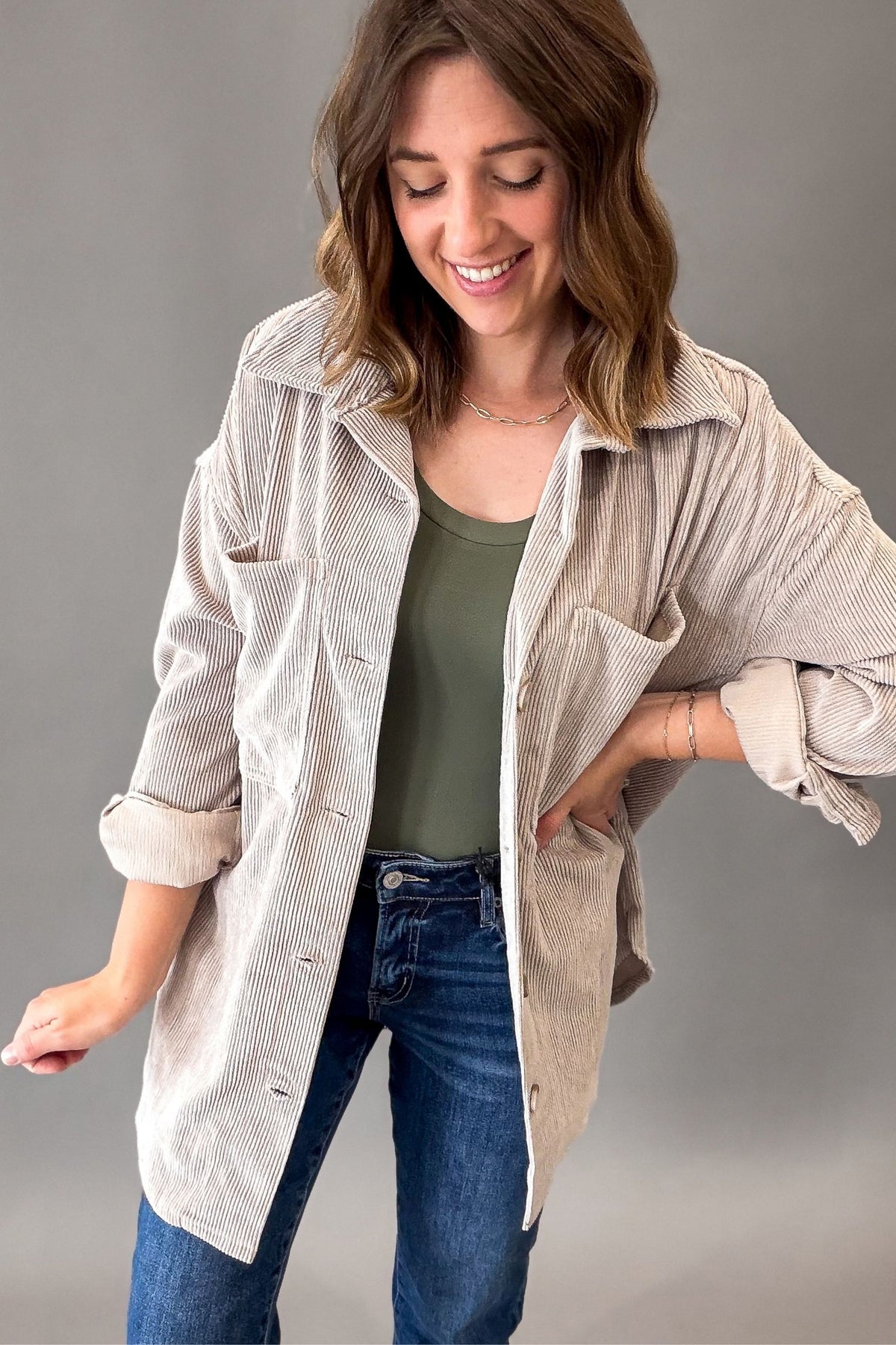 A woman wearing a light beige corduroy button-up shirt over an olive green tank top and blue jeans, posing against a neutral gray studio background.
