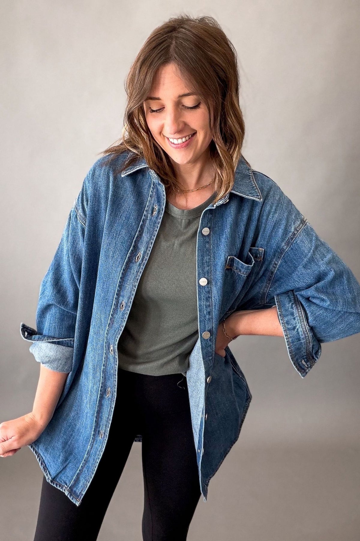 A woman wearing an oversized blue denim shirt over a muted green t-shirt and black pants, posing against a neutral gray studio background.