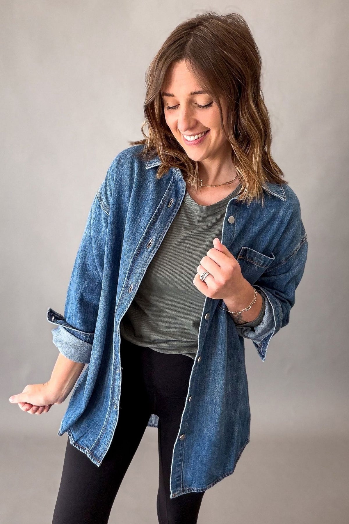 A woman wearing an oversized blue denim button-up shirt over a gray t-shirt and black leggings, posing against a neutral gray studio background.