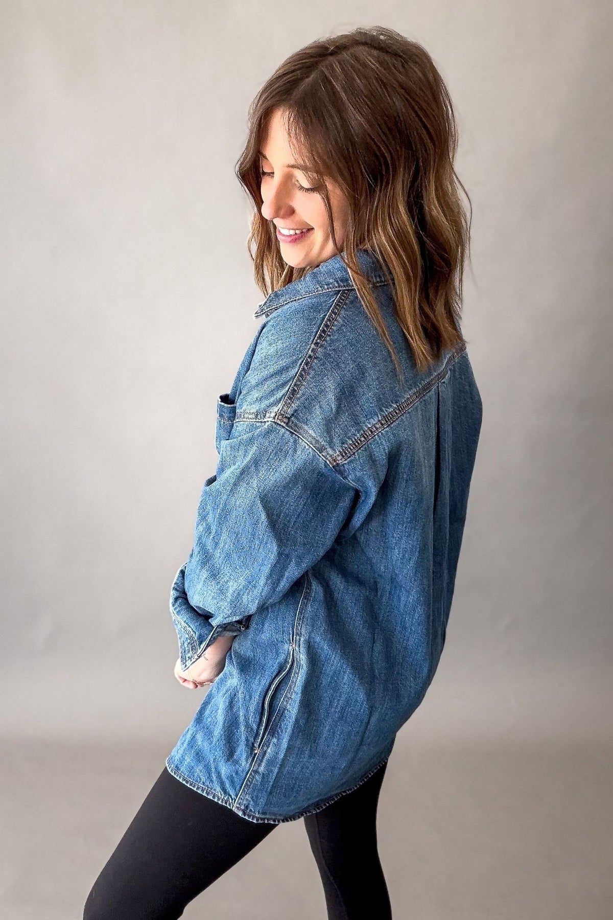 A woman wearing an oversized blue denim jacket and black leggings, posing in profile against a neutral gray studio background.