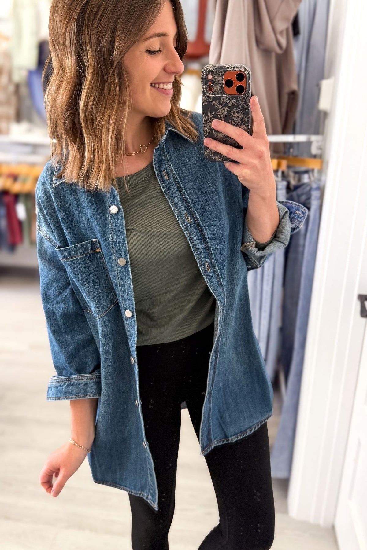 A woman takes a mirror selfie in a clothing store, wearing a blue denim button-up shirt over an olive green t-shirt and black pants.