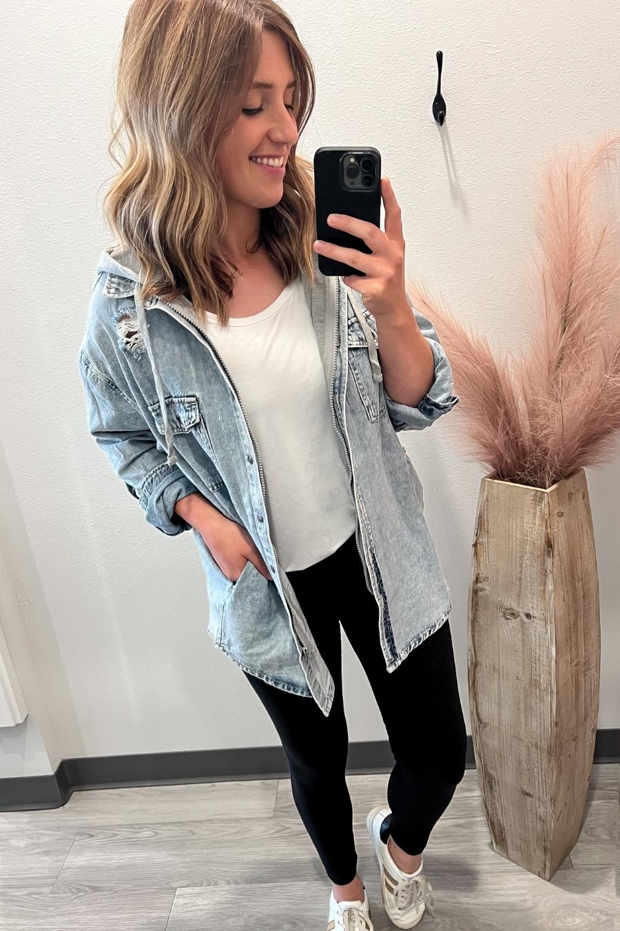 Woman taking a selfie in a casual outfit with a denim jacket and white shirt, standing next to a wooden vase with pink dried plants.